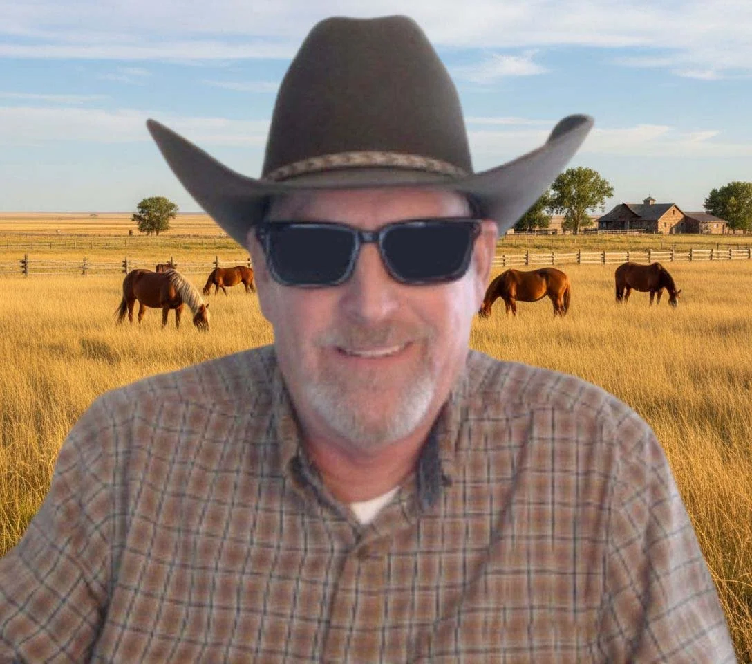 Man wearing cowboy hat and sunglasses smiling in front of a rural farm background with horses, farm buildings, trees, and wooden fences.