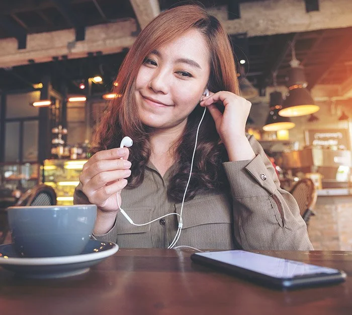 A young woman in a coffee shop prepares to receive a telehealth appointment with The Dietitian's Office using her phone and earbuds.