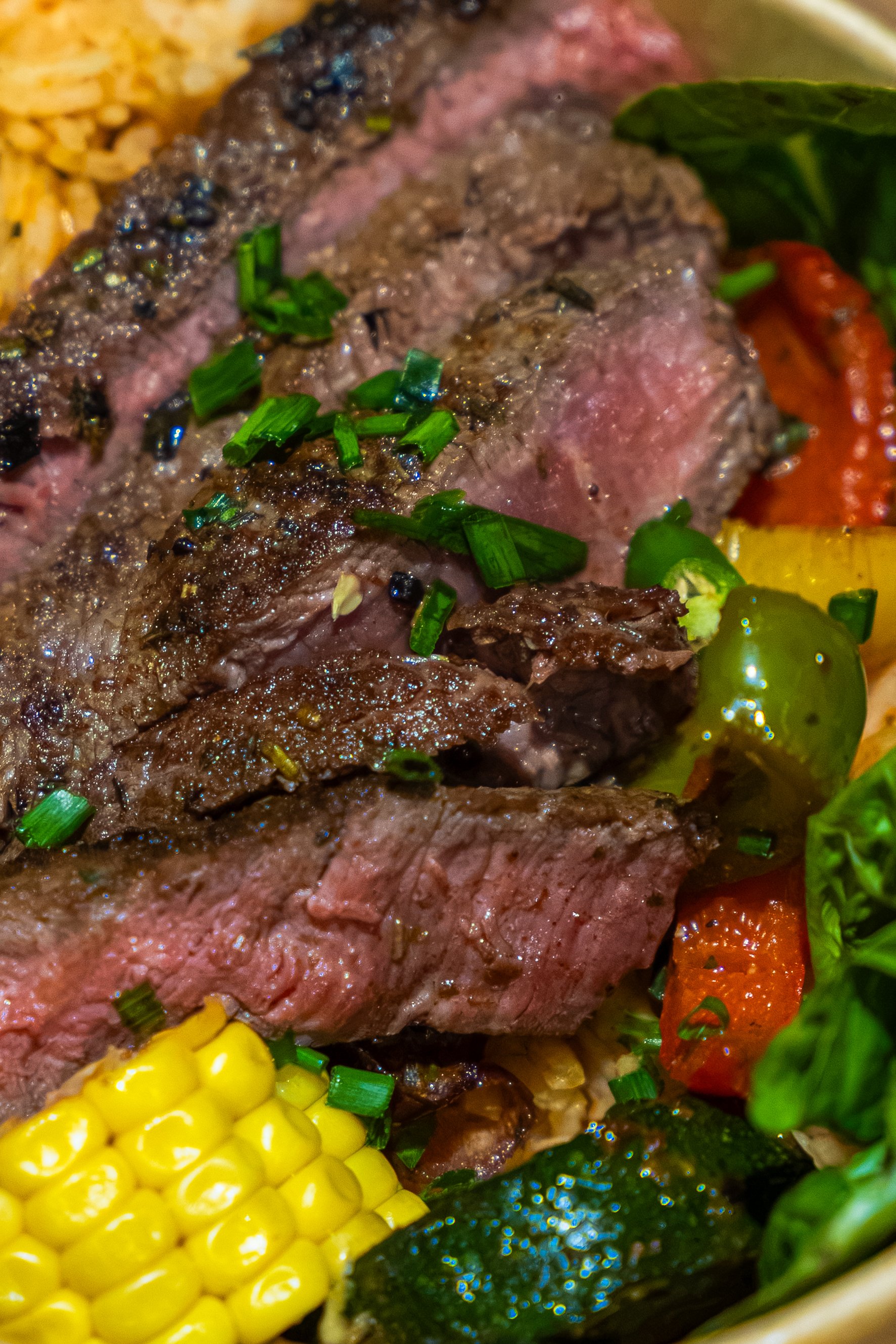 Close-up of a cooked steak topped with chopped green herbs, served with yellow corn on the cob, cherry tomatoes, and leafy greens.