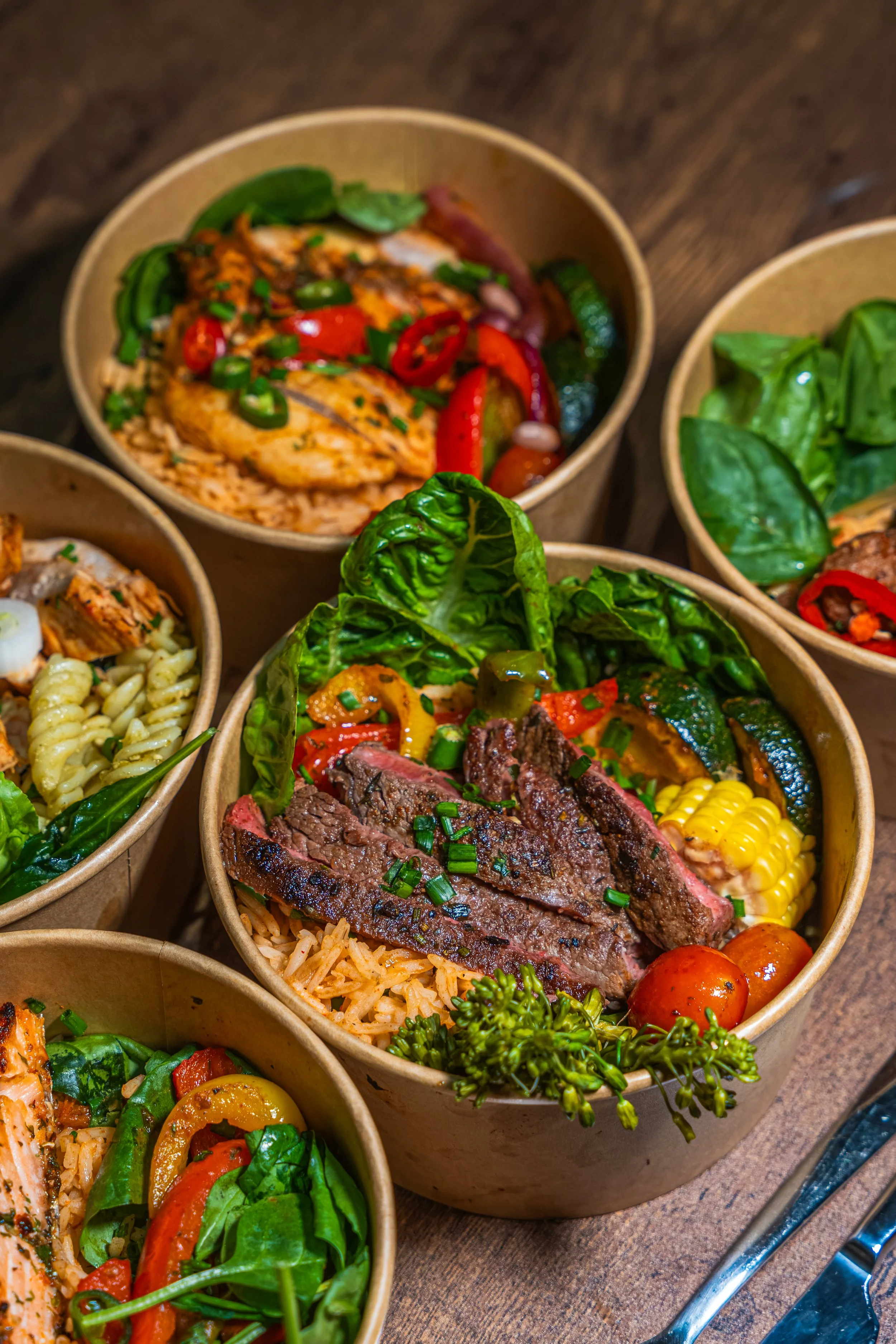 Assorted bowls of healthy salads with grilled meats, vegetables, pasta, and herbs on a wooden table.