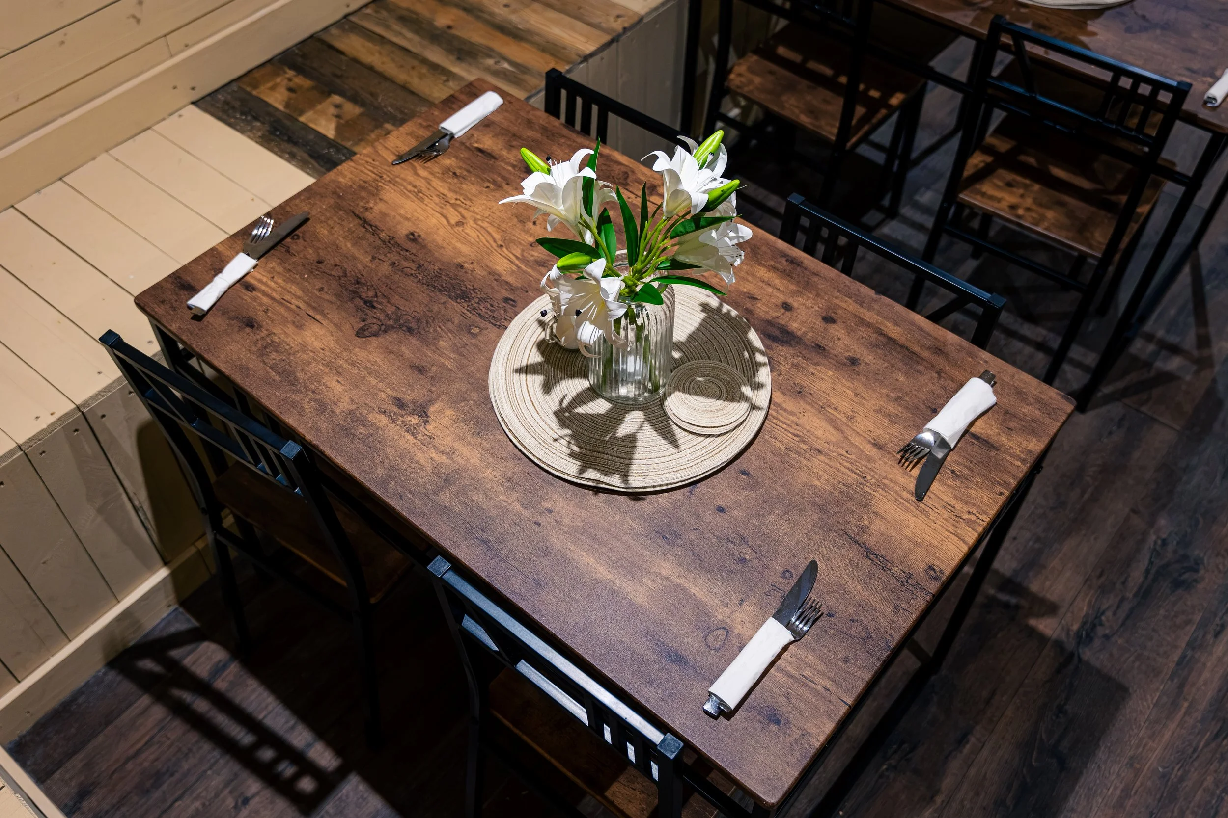 A wooden dining table with a vase of white lilies, a round placemat, and cutlery wrapped in napkins at each seat. The table is in a rustic-style space with wooden flooring.