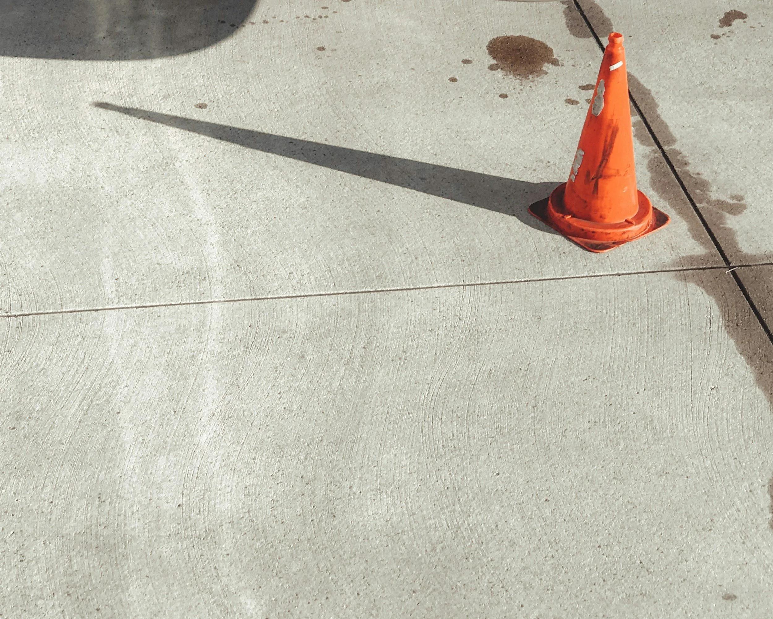 An orange traffic cone on a concrete sidewalk with a black cord running across it. There are wet spots on the ground, and the cone casts a shadow.