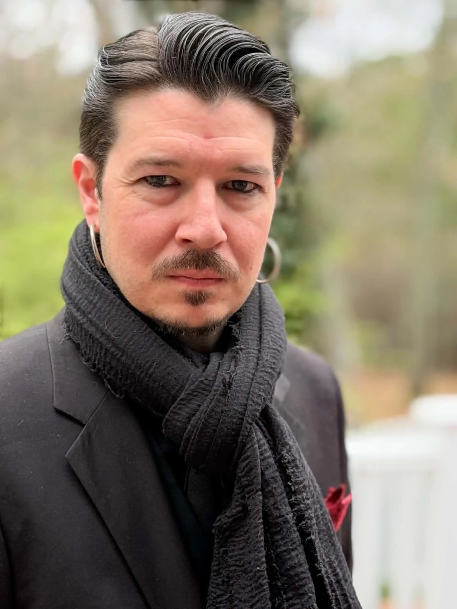 A man with dark, slicked-back hair, blue eyes, a goatee, on an outdoor setting with blurred greenery background, wearing dark clothing and a gray scarf.