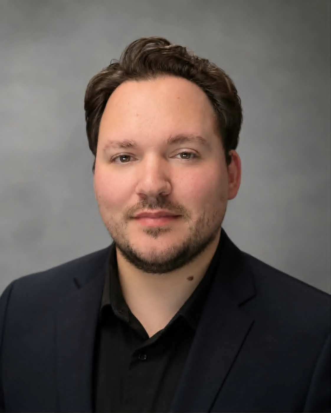 Headshot of a man with dark brown hair, light skin, wearing a black suit jacket and shirt, against a plain gray background.