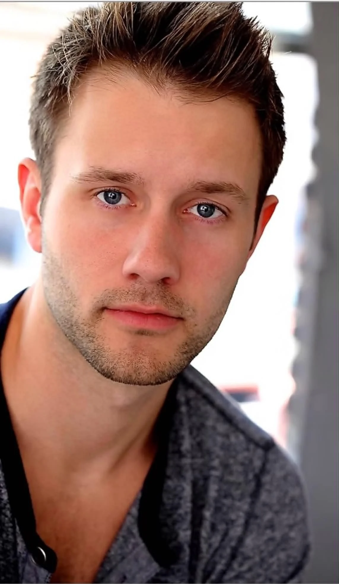 Close-up portrait of a young man with blue eyes, light facial hair, and short dark hair with highlighted tips, wearing a gray jacket over a black shirt, in a well-lit indoor setting.