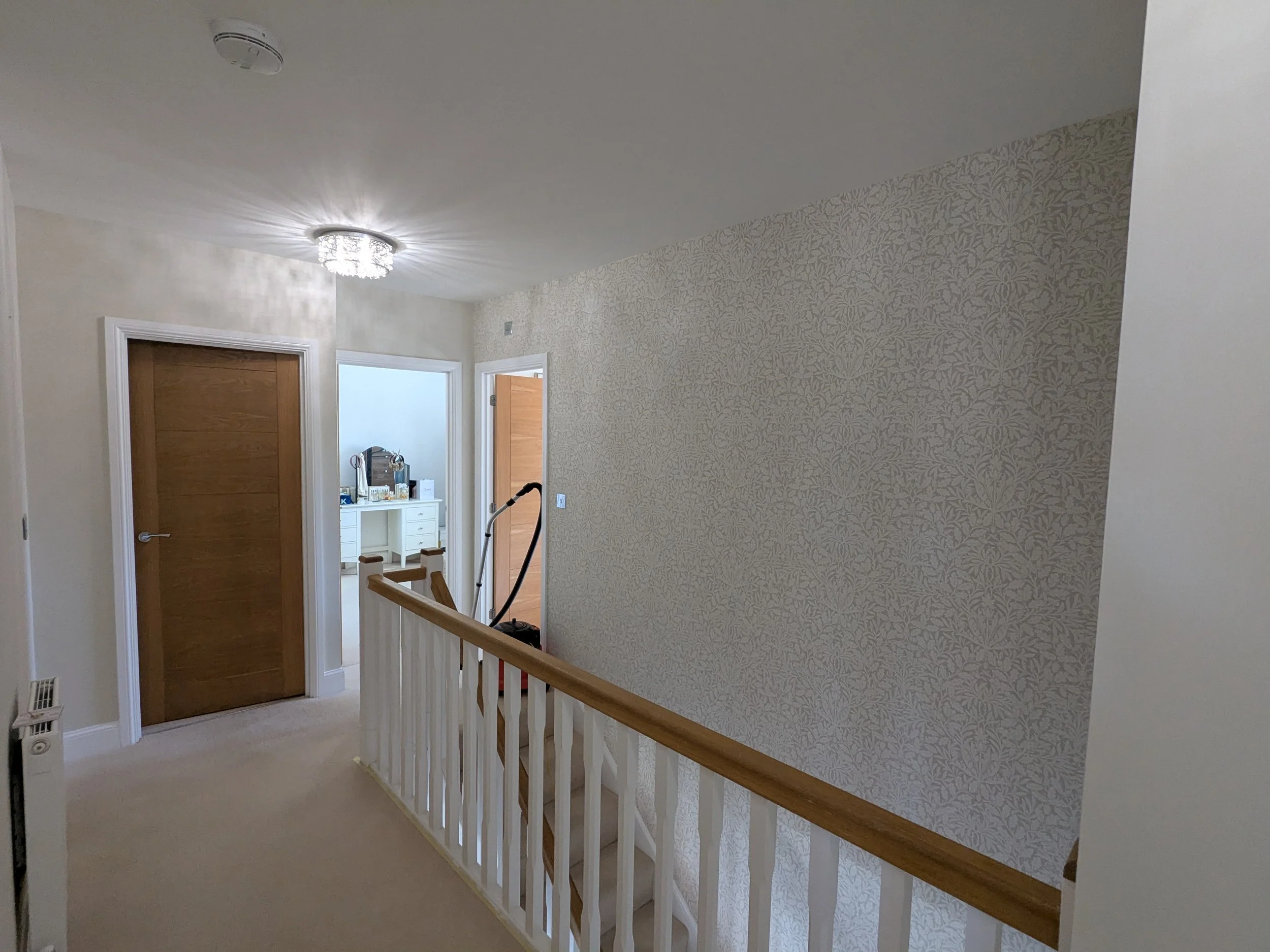 Interior view of a staircase landing with beige carpet, a wooden handrail, and walls decorated with light patterned wallpaper. There are two doors, one closed wooden door and one open door leading to a room with a white dresser. A ceiling light fixtu