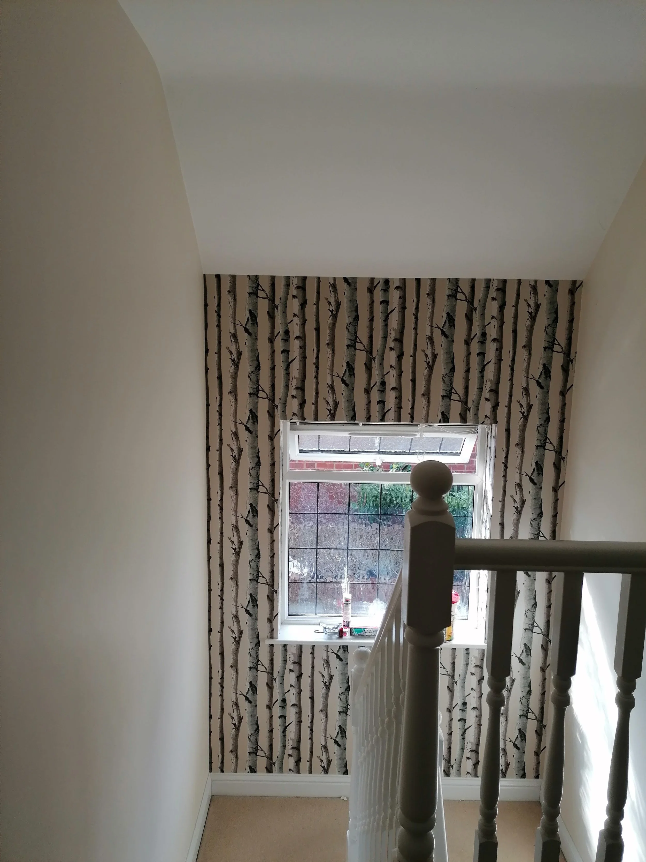 Interior view from the top of a staircase showing a window with birch tree wallpaper behind it, a white radiator below the window, and a white spindled staircase railing in the foreground.