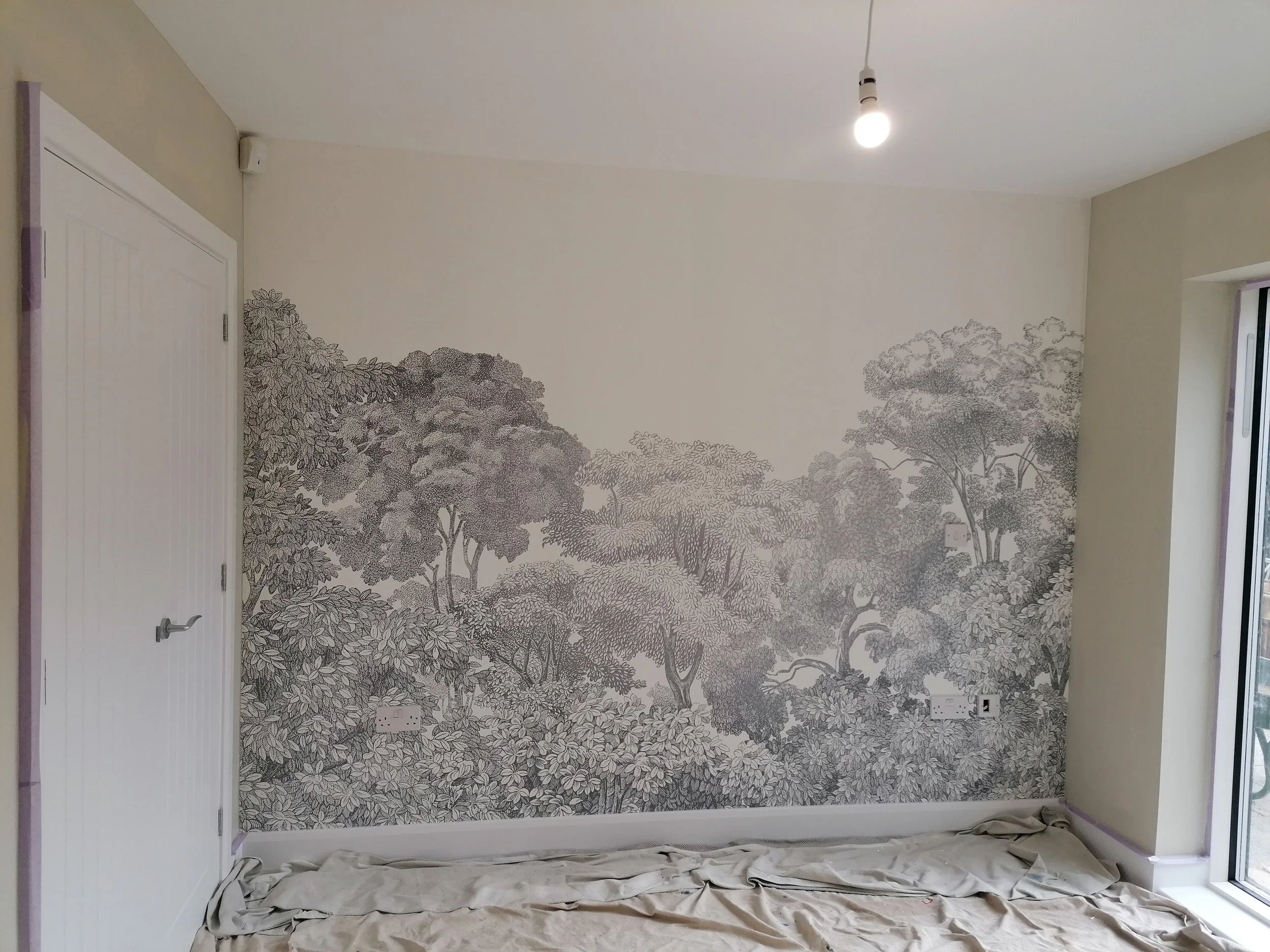 Interior of a room with a decorative wallpaper of trees on the wall, white ceiling, and a hanging light bulb. The floor is covered with a protective sheet, and there's a partially visible door on the left and a window on the right.