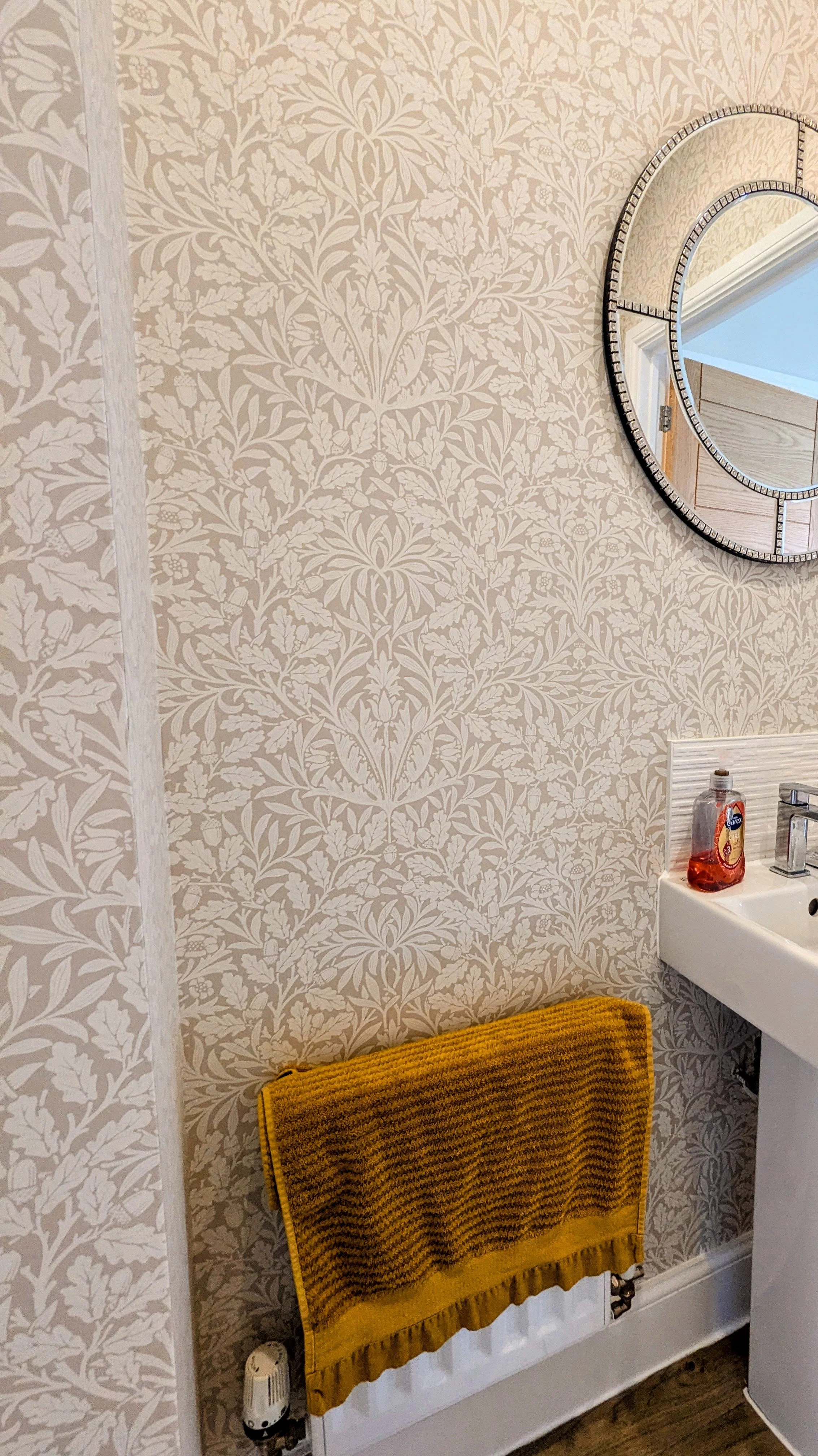 Bathroom with beige wallpaper featuring a floral pattern, a round mirror with a decorative frame hanging above a white sink, a bottle of hand soap on the sink, a radiador with a yellow towel hanging on it, and a wooden floor.