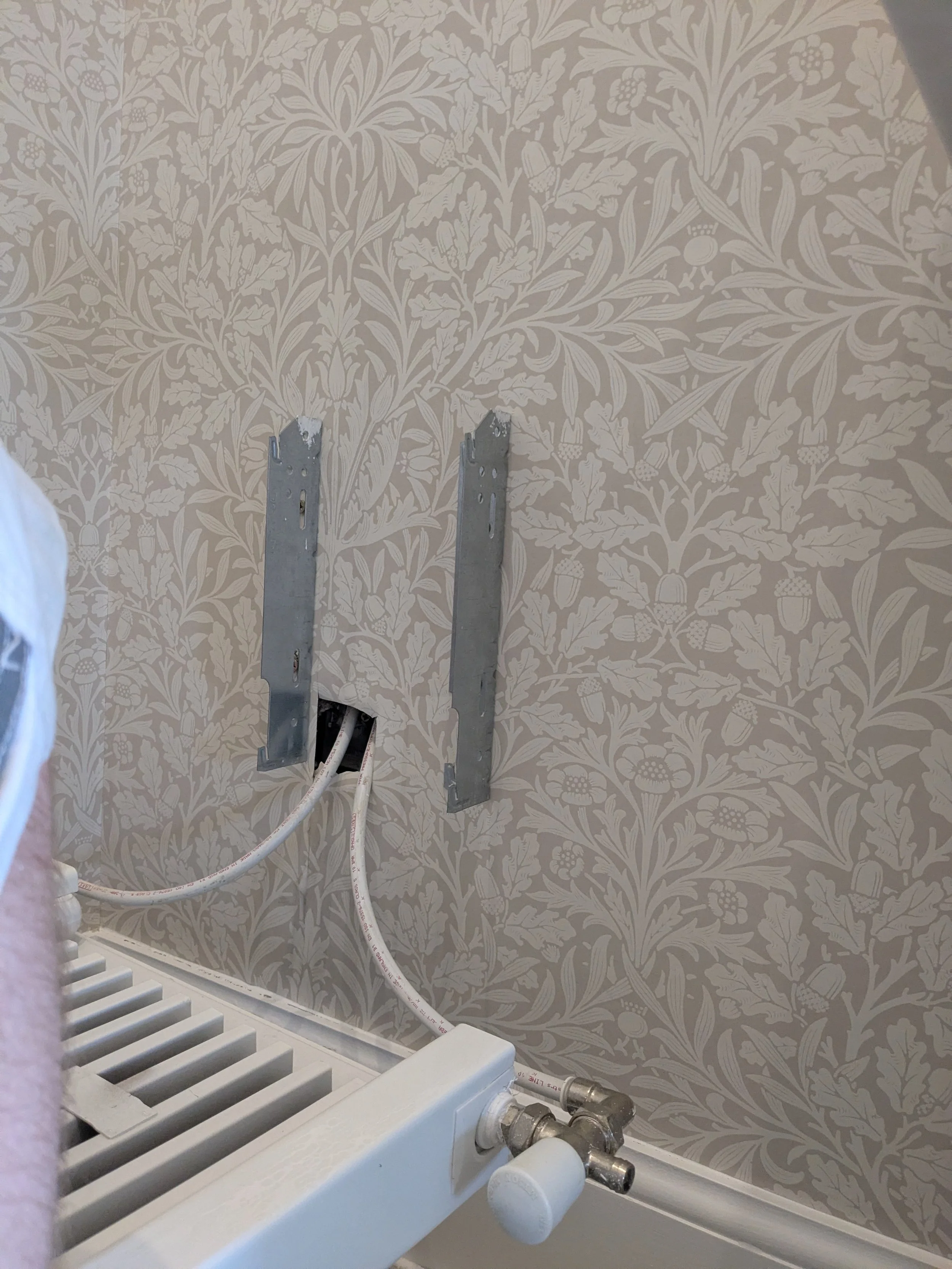 A partially installed radiator or heater with exposed pipes, mounted on a wall with beige floral wallpaper, and two metal brackets above it.