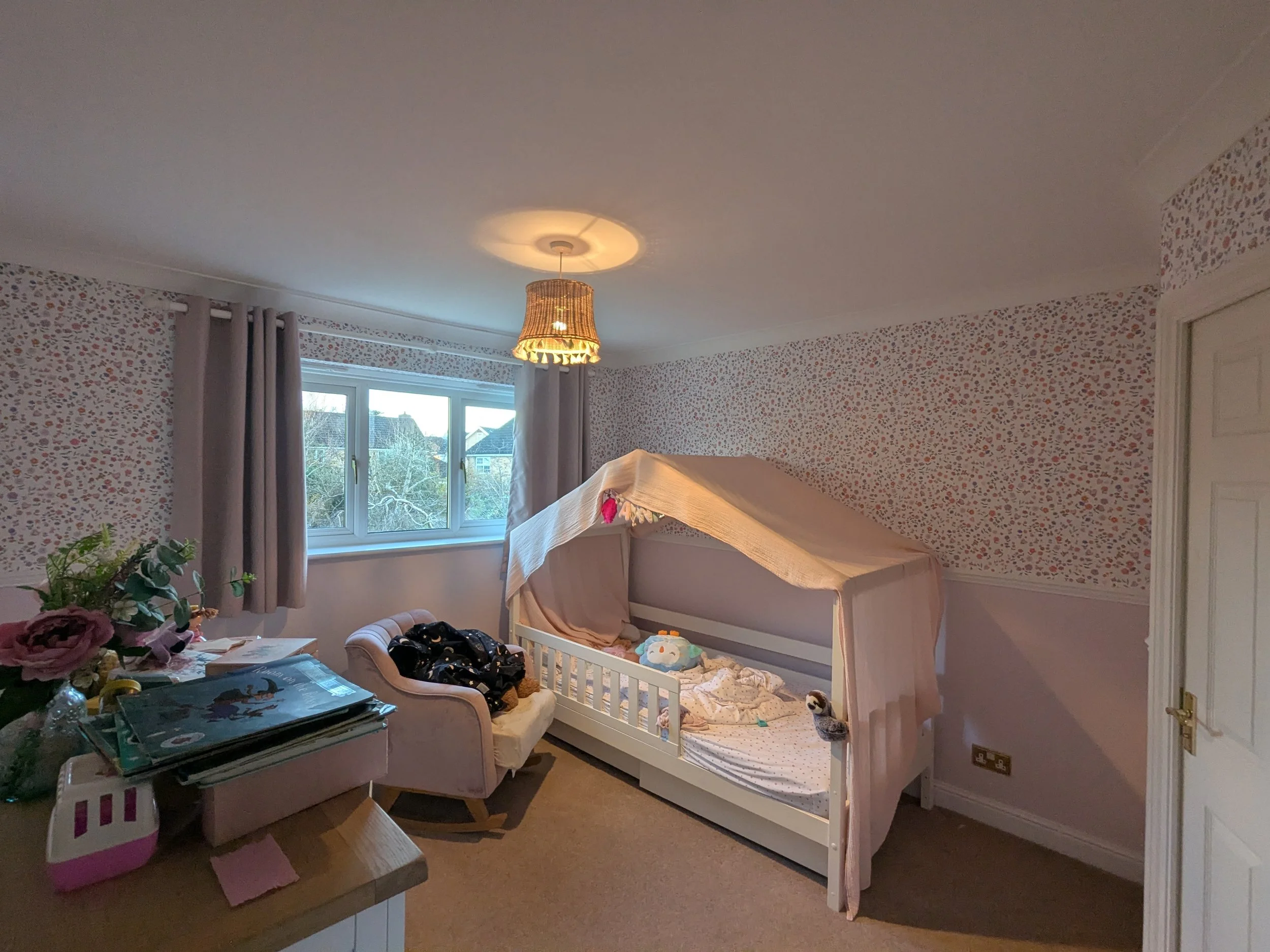 A bedroom with floral wallpaper, a window with grey curtains, a baby crib with bedding and a stuffed animal, a beige armchair with a black bag on it, a table with piles of books and flowers, and a ceiling light with a lampshade.