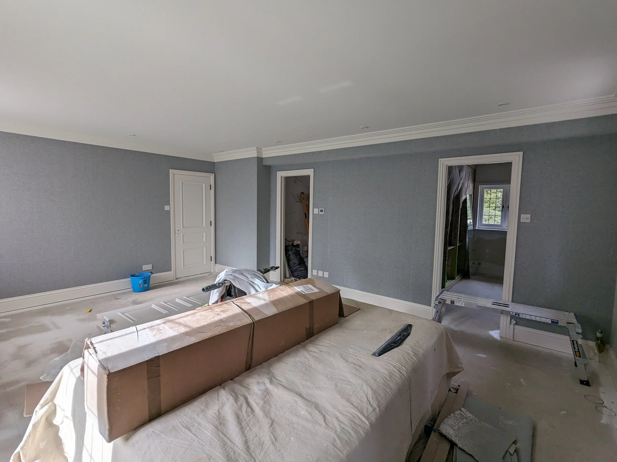 Living room under renovation with construction materials and tools, including a large cardboard box, a dust sheet, and a window with sunlight shining through.