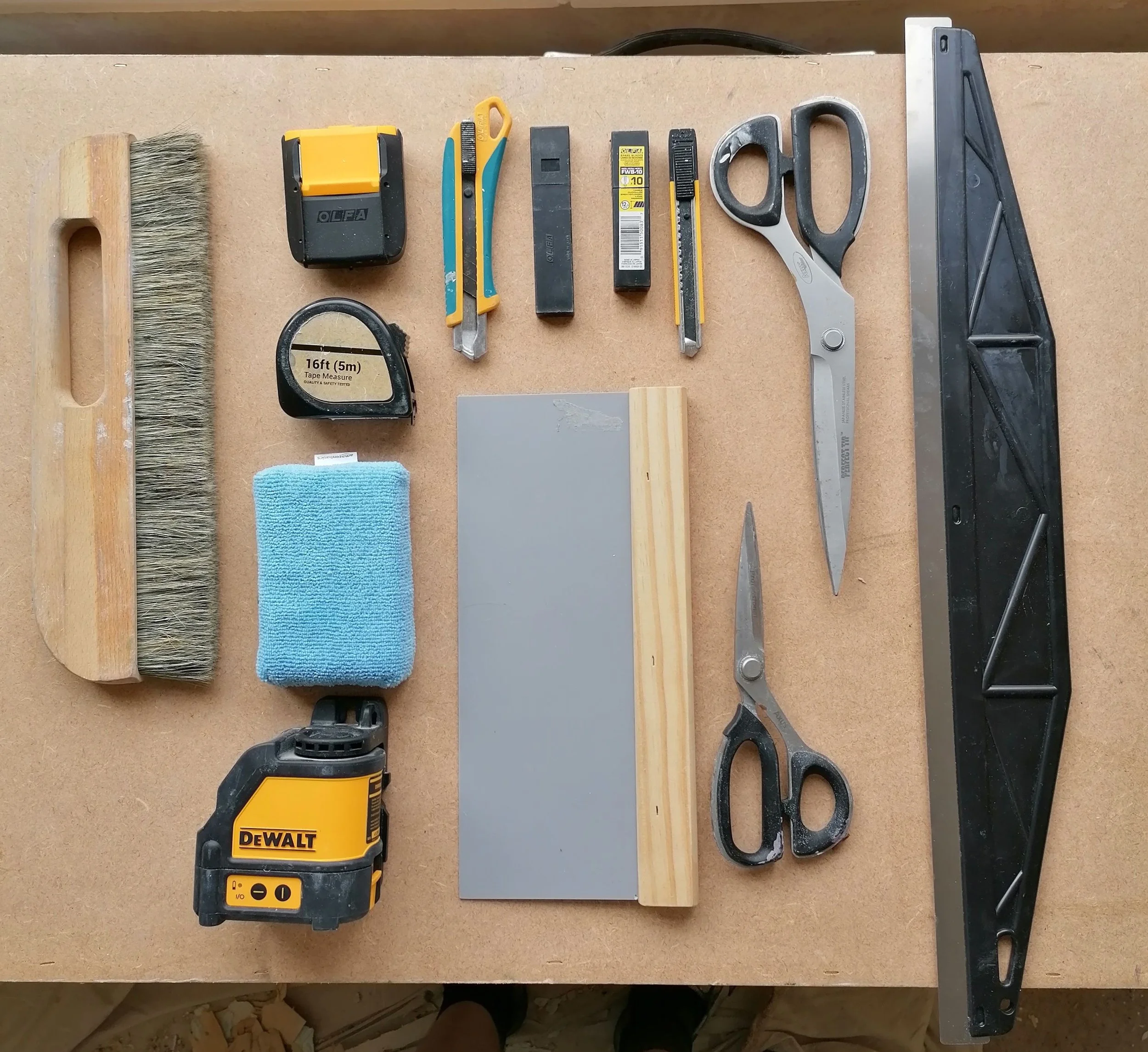 Tools on a workbench including a paperhanging brush, tape measure, utility knife, black marker, shears, square, circular saw, and a handsaw. Wooden block, gray metal sheet, and a blue cloth are also visible.