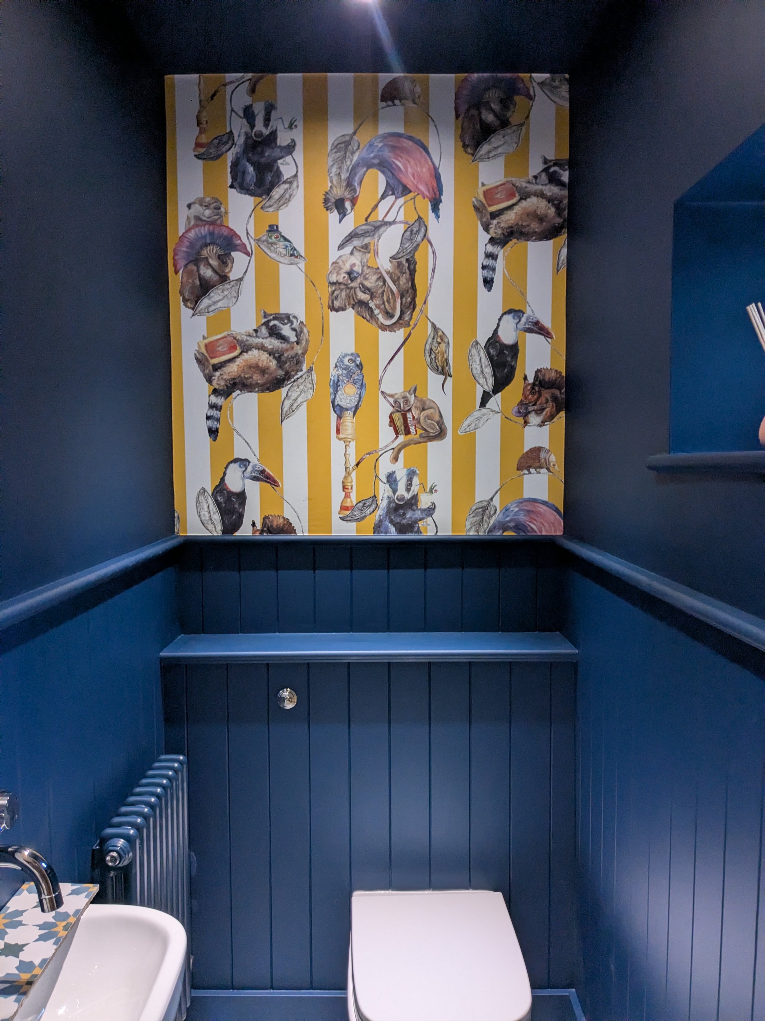 The image shows a small, dark-colored room with a blue wooden wall and a yellow and white striped artwork featuring various animals, including birds, raccoons, and other creatures. There is a white sink with a patterned counter on the left and a small white table or countertop at the bottom. A metallic handrail is visible on the left side.