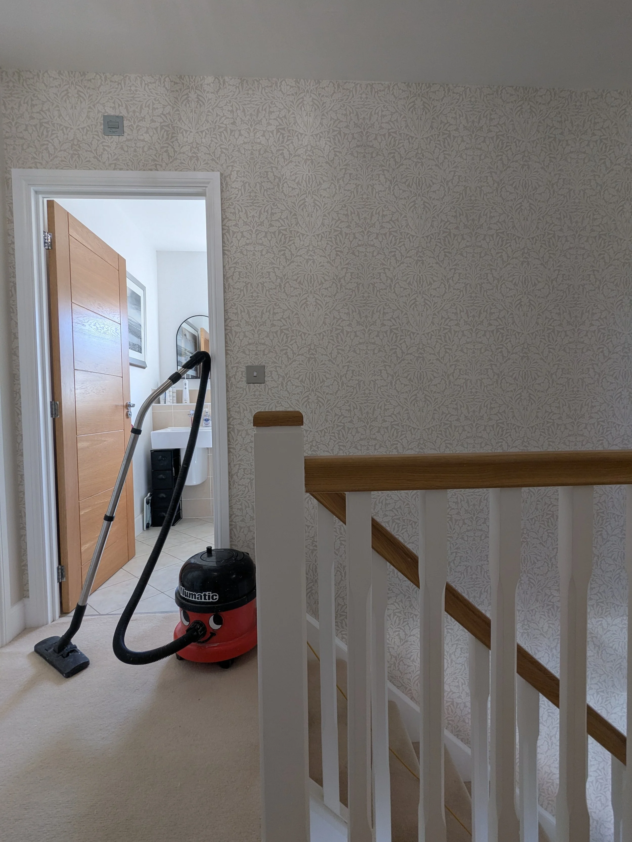 Vacuum cleaner on carpeted floor near a staircase with wooden railing, with bathroom visible through open door in the background.