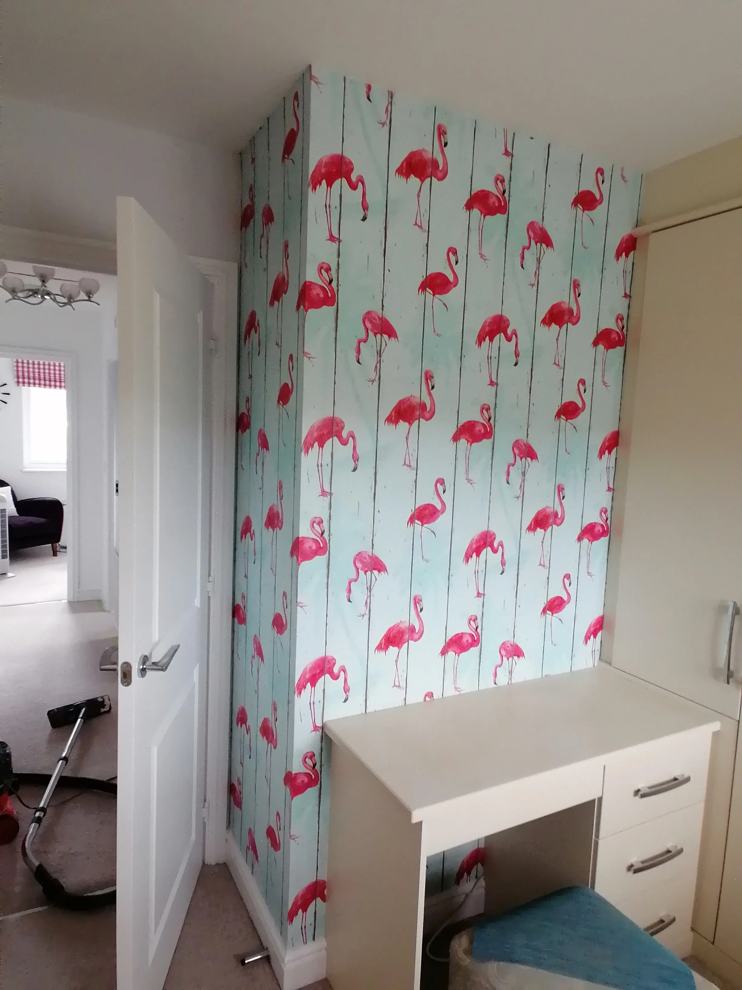 Interior view of a room with a wall decorated with pink flamingo wallpaper, a white cabinet, a desk, and a partially open door leading to another room.