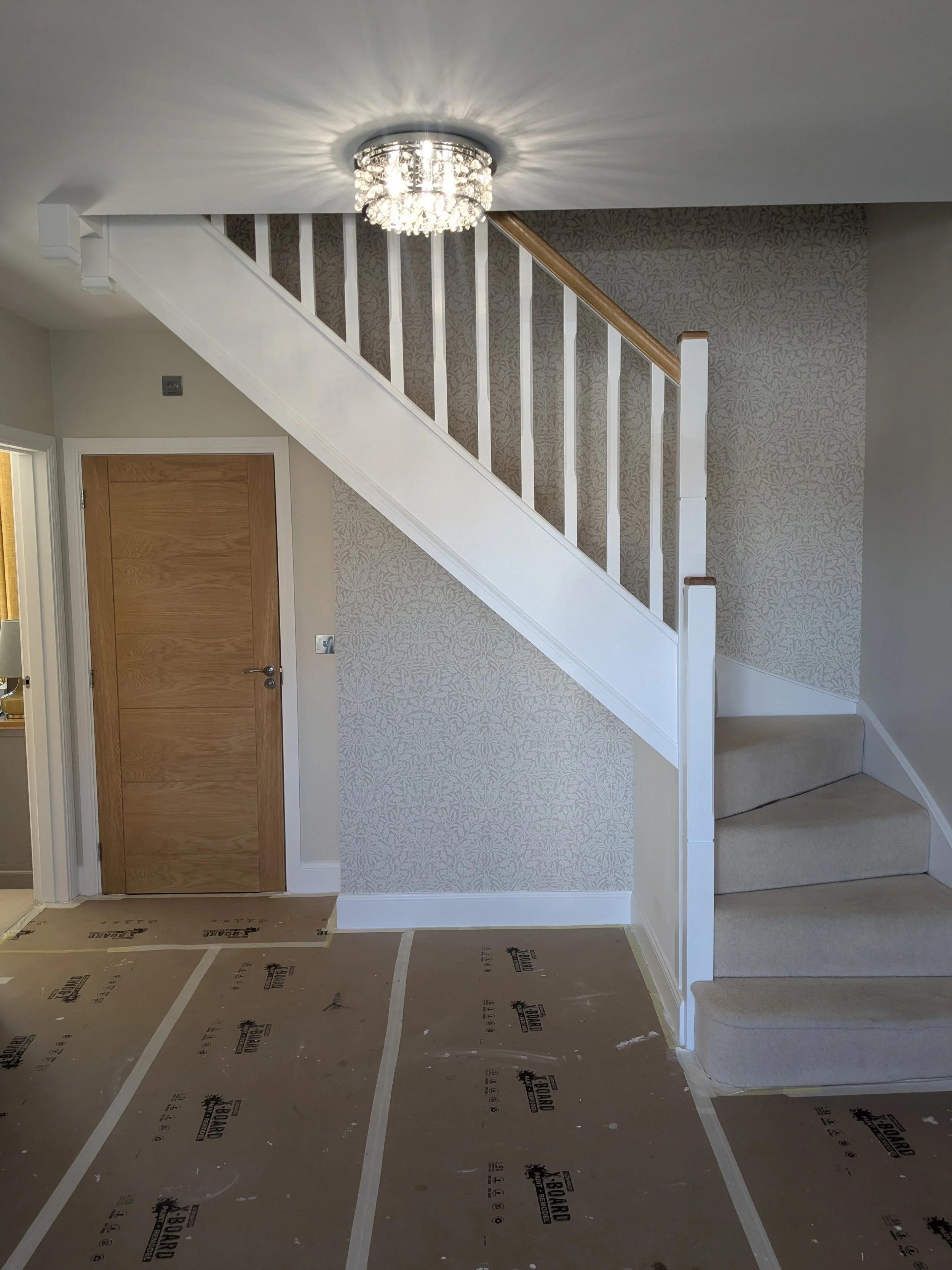 Interior view of a staircase with a chandelier, a closed wooden door, wallpapered wall, and flooring covered with protective material.