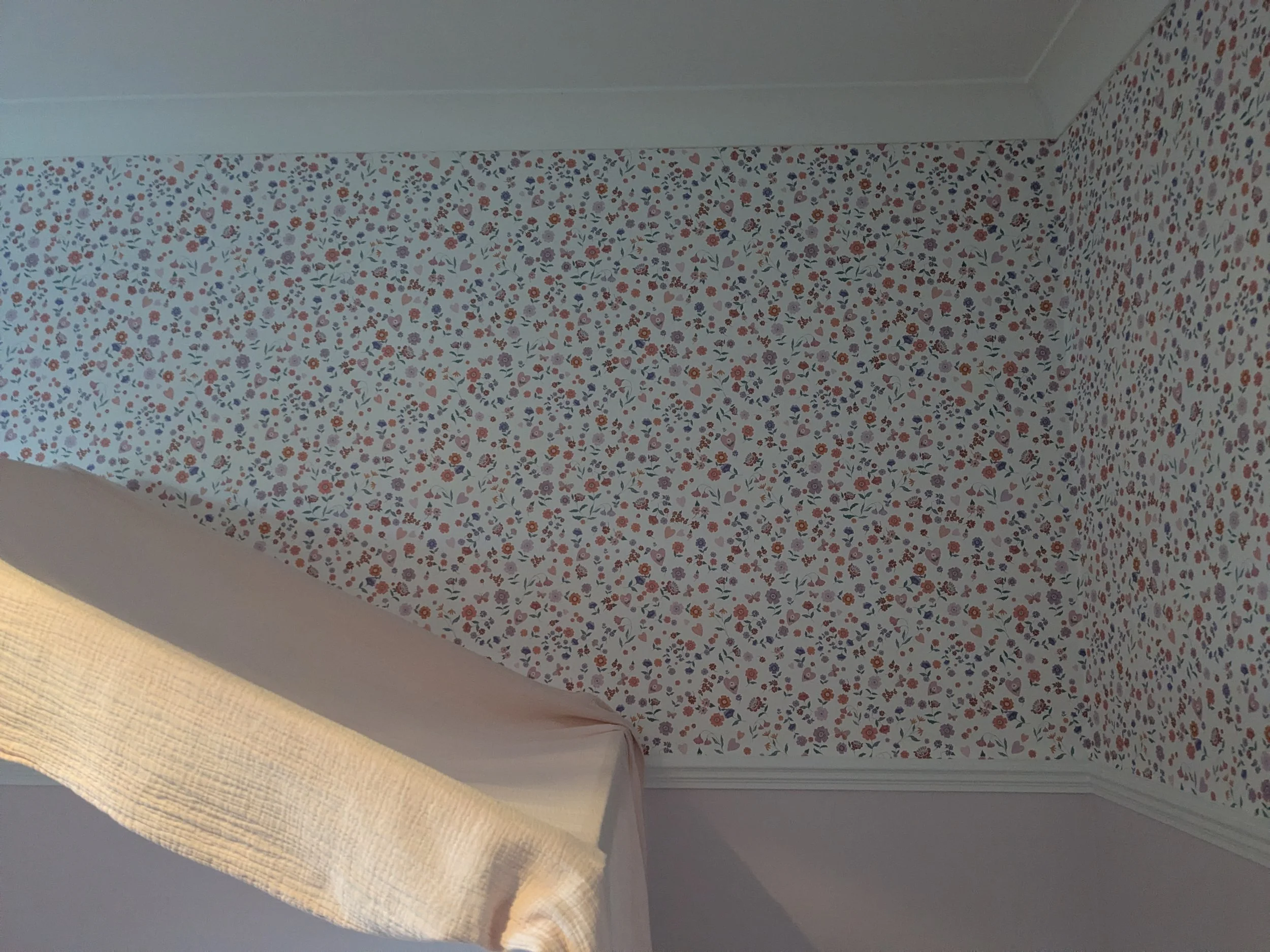 Close-up image of a wall covered in floral wallpaper with a corner, showing a bed with a beige blanket in the lower left corner.