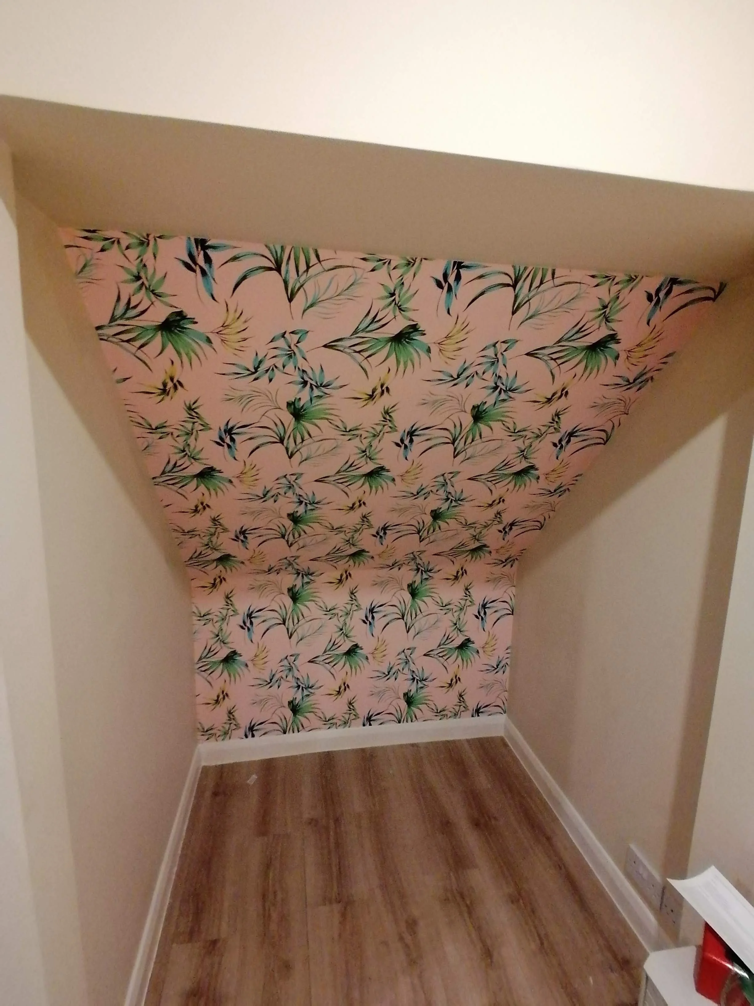 A small corner of a room with a window covered by pink floral patterned curtains, wooden flooring, and beige walls.