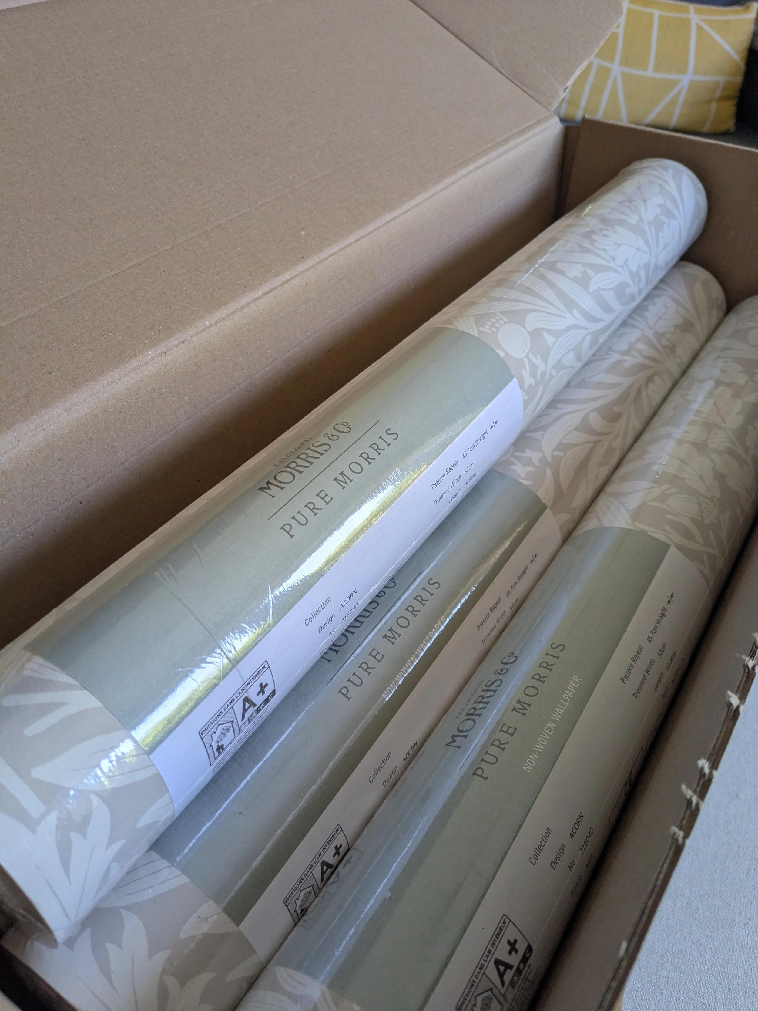 Line of wallpaper rolls inside a cardboard box, labeled 'Pure Morris' with decorative patterns on the rolls.