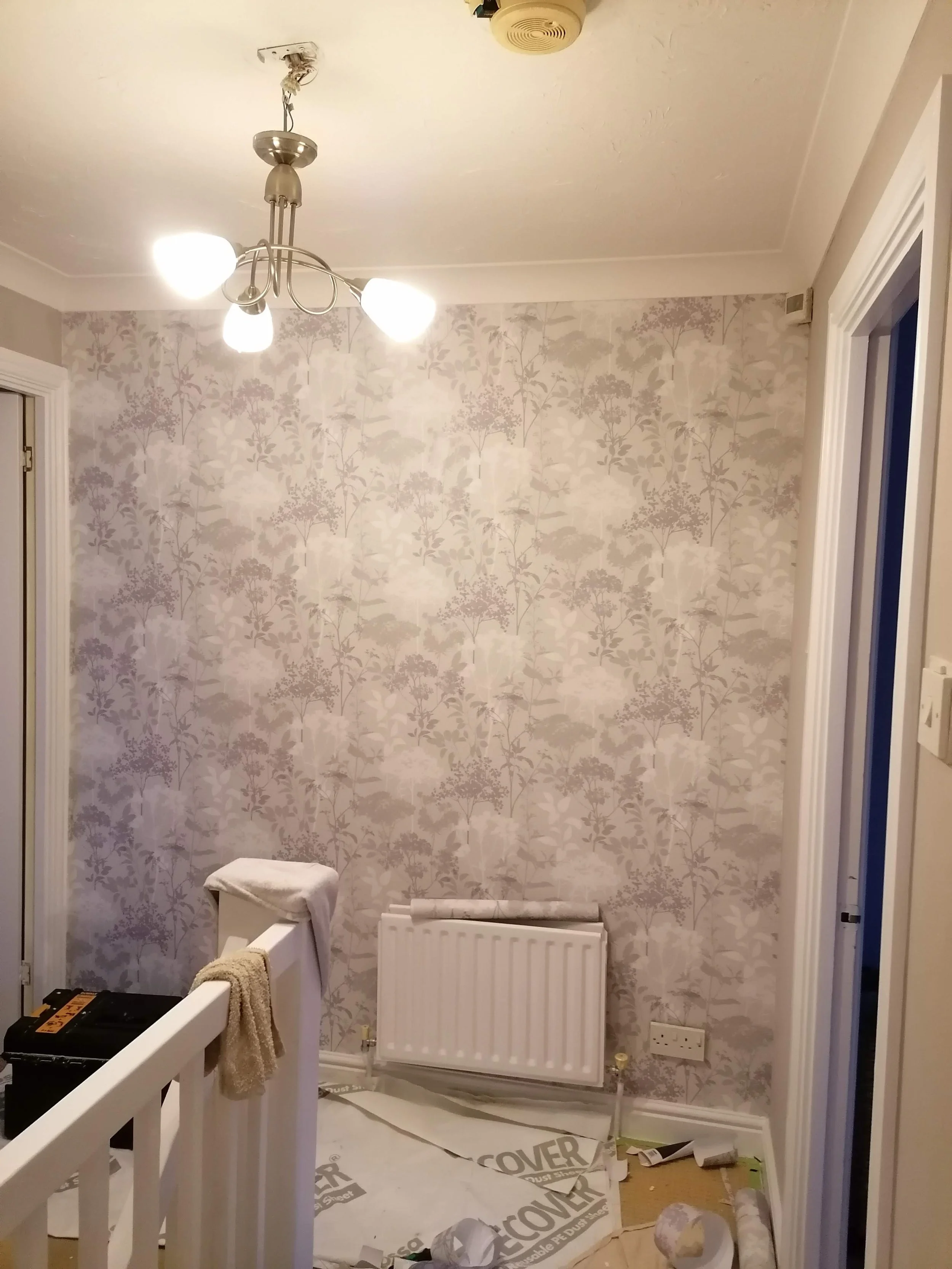A partially renovated room with wallpapered walls, a ceiling light fixture, a radiator, and construction materials on the floor. There's a small stool covered with a cloth and a white railing in the foreground.