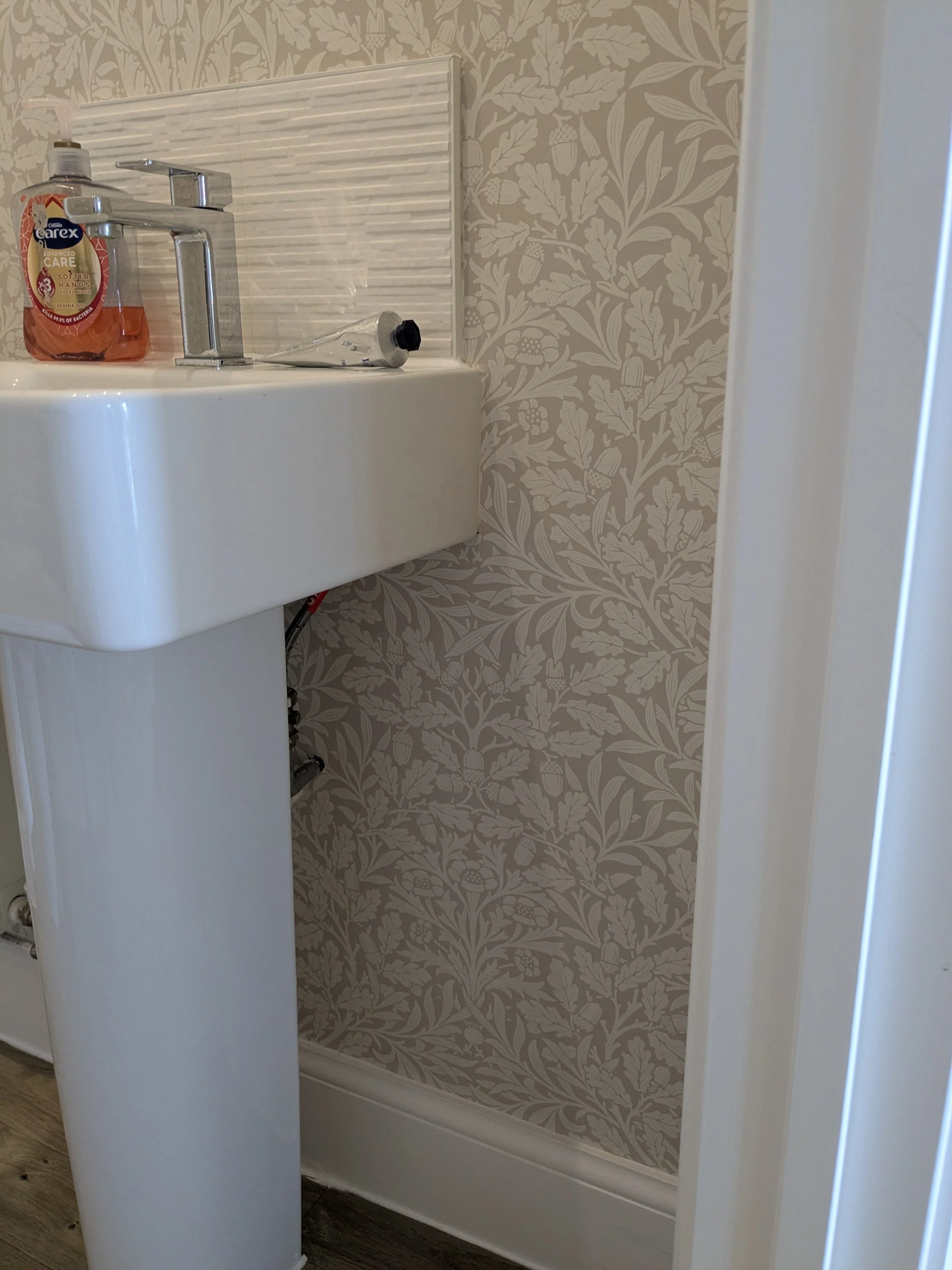 A bathroom corner with a white sink, a chrome faucet, a bottle of Carex hand soap, and a tube of hand cream. The wall has patterned wallpaper with leaves and acorns, and there is a partial view of a door frame.