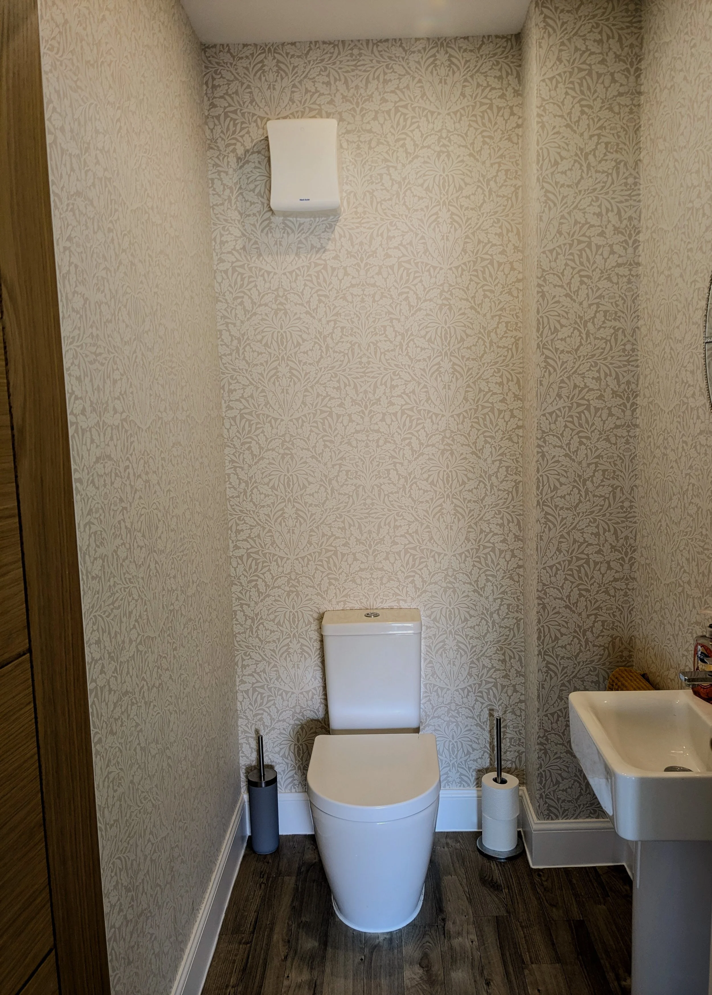 A small bathroom with a wall-mounted white toilet, a gray toilet brush holder, a toilet paper roll, a wall-mounted white hand dryer, a small sink with a red and black soap dispenser, and wallpaper with a beige floral pattern. The floor is dark wood.