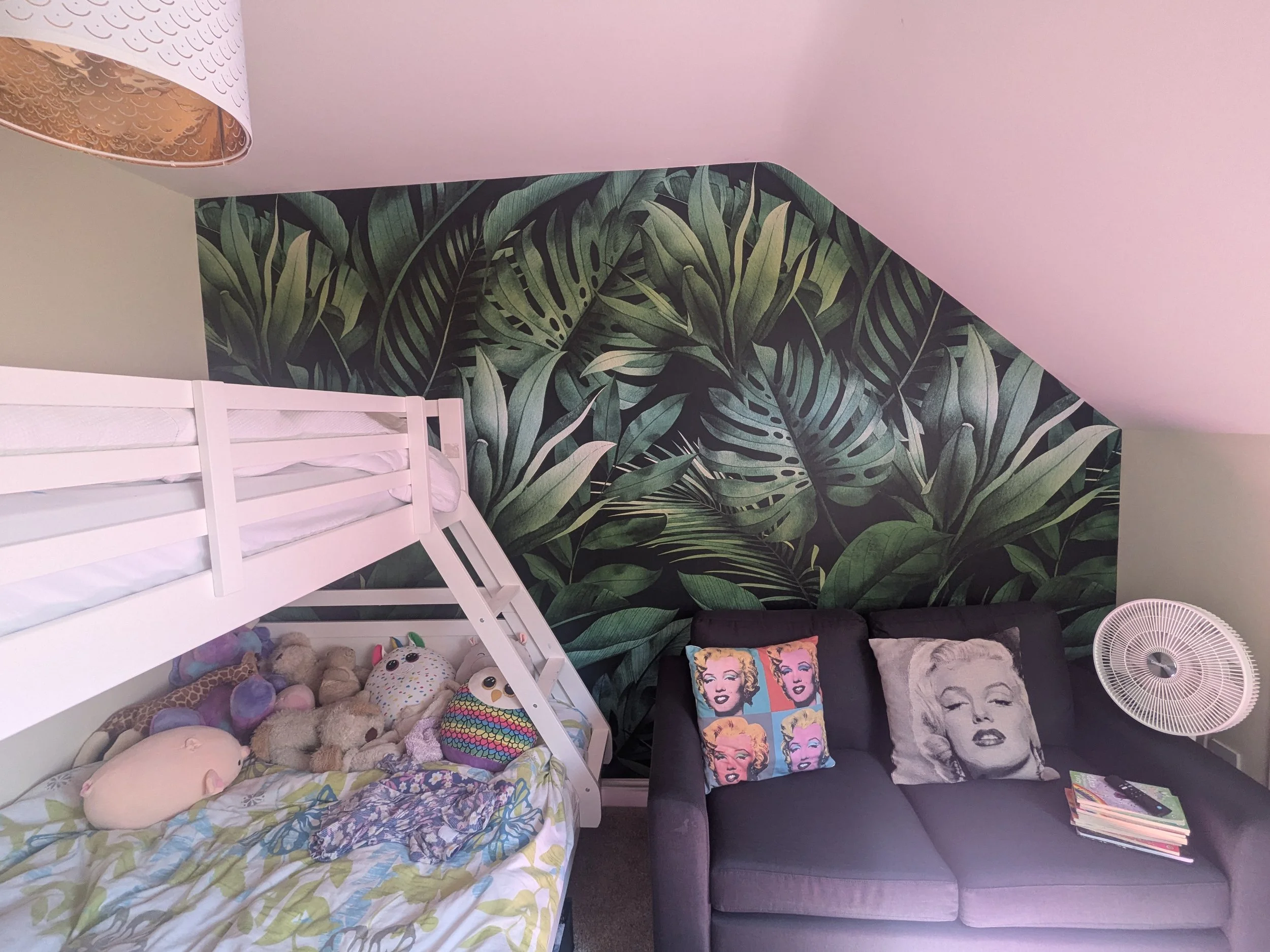 A bedroom with a lush green tropical leaf wallpaper on the back wall, a white bunk bed with stuffed animals, pillows, and bedding on the bottom bunk, a black sofa with decorative pillows featuring Marilyn Monroe, a small white fan, and a stack of mag
