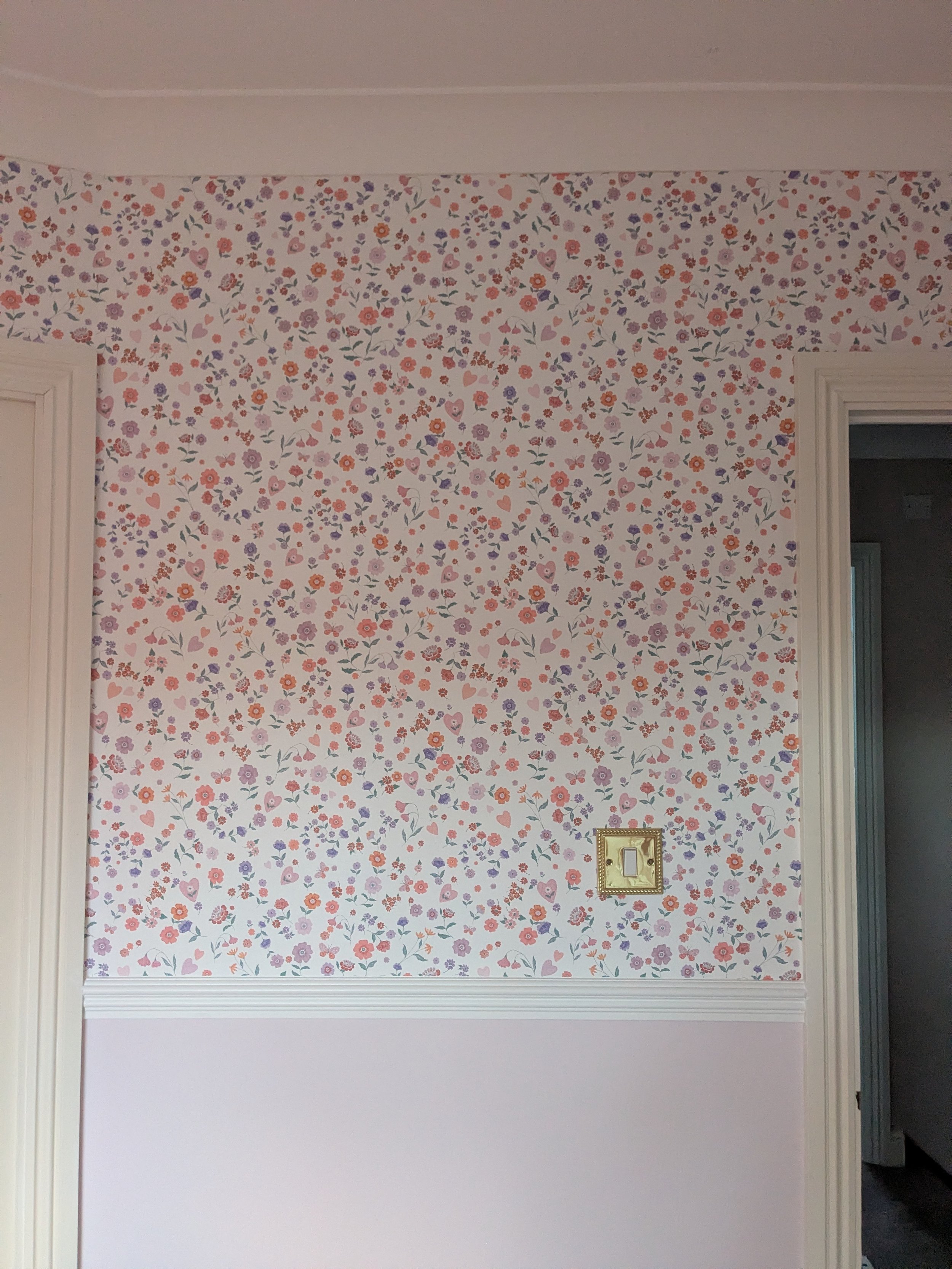 A wall with floral wallpaper featuring small pink, purple, and orange flowers with green leaves, and a light switch, with a doorway on the right side.