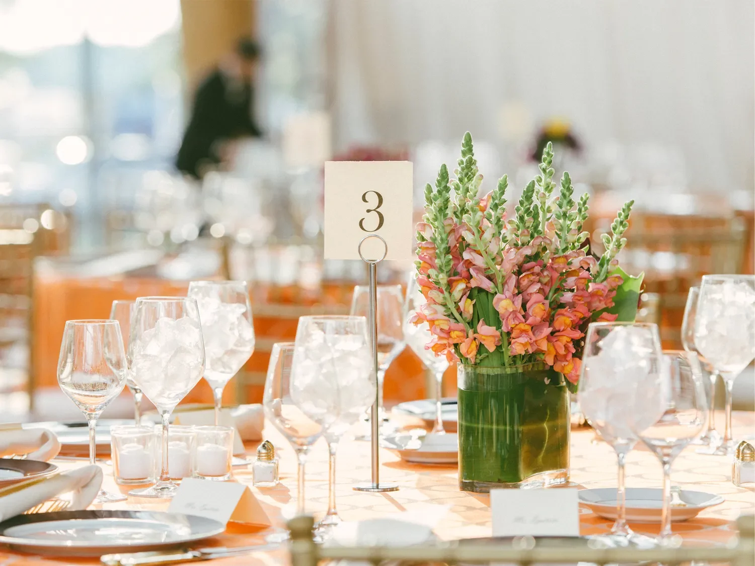 A table setting at a wedding or event with a floral centerpiece, wine glasses, and a table number 3 sign.