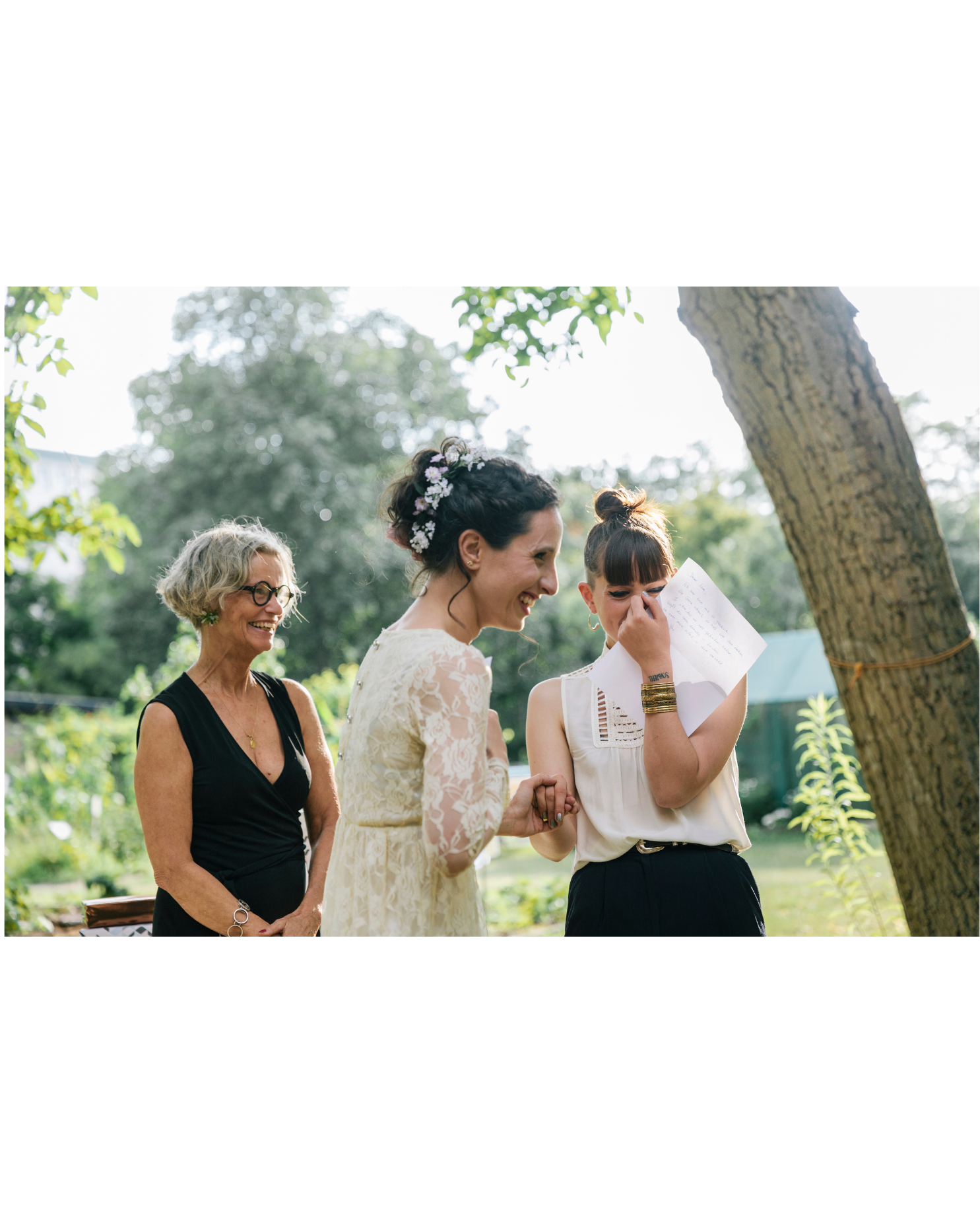 Four women outdoors, gathered around and smiling, one woman holding a piece of paper, setting appears to be a garden or park, during a sunny day.