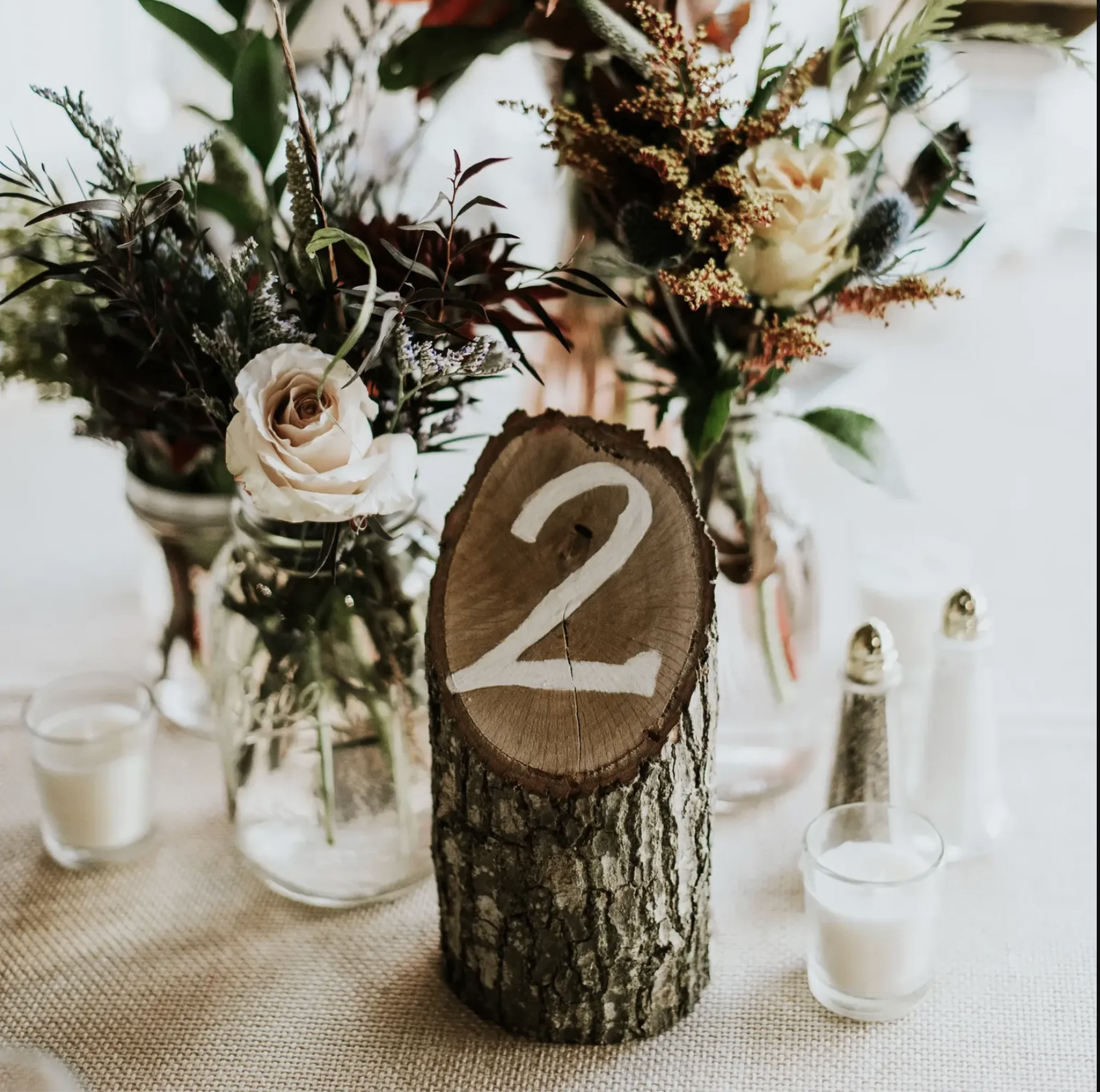A floral centerpiece with white roses and various greenery, with a wooden log marked with the number 2, surrounded by small candles in glass holders on a beige tablecloth.