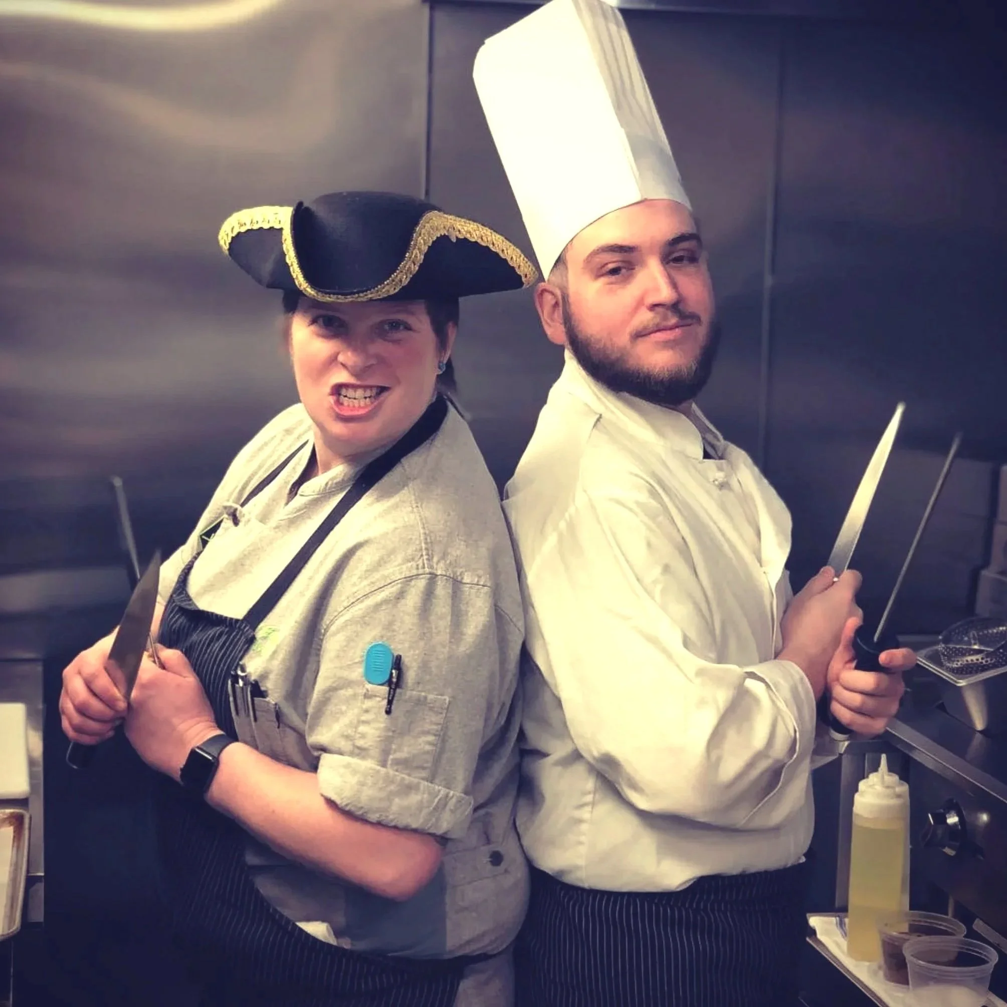 Two chefs wearing tall white hats and chef coats, standing side by side in a kitchen.