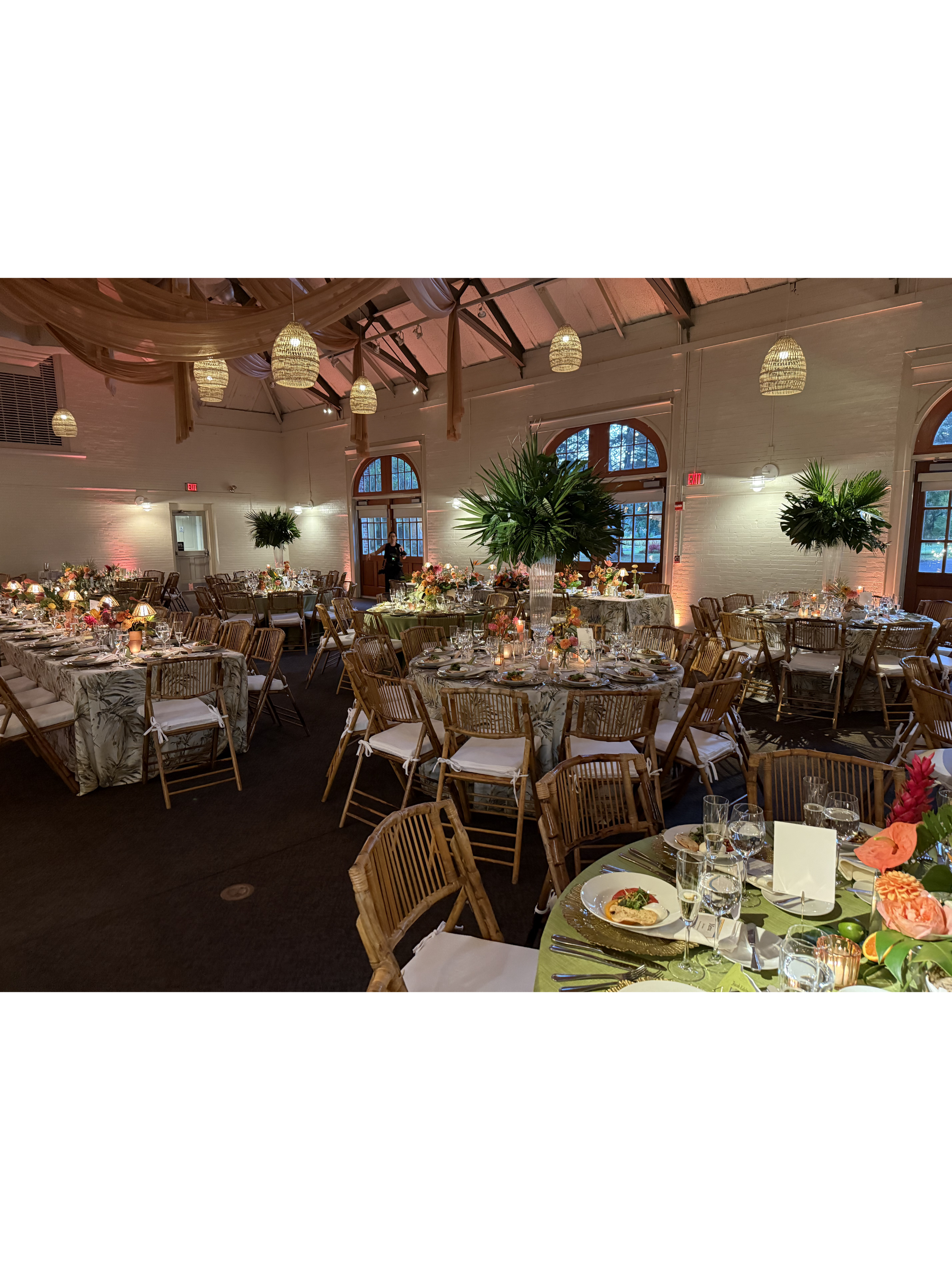 Elegant reception hall decorated with round tables covered in floral tablecloths, set with dinnerware and glassware, surrounded by wicker chairs, with large green plants in the center, warm lighting, and high vaulted ceiling with hanging woven lamps.