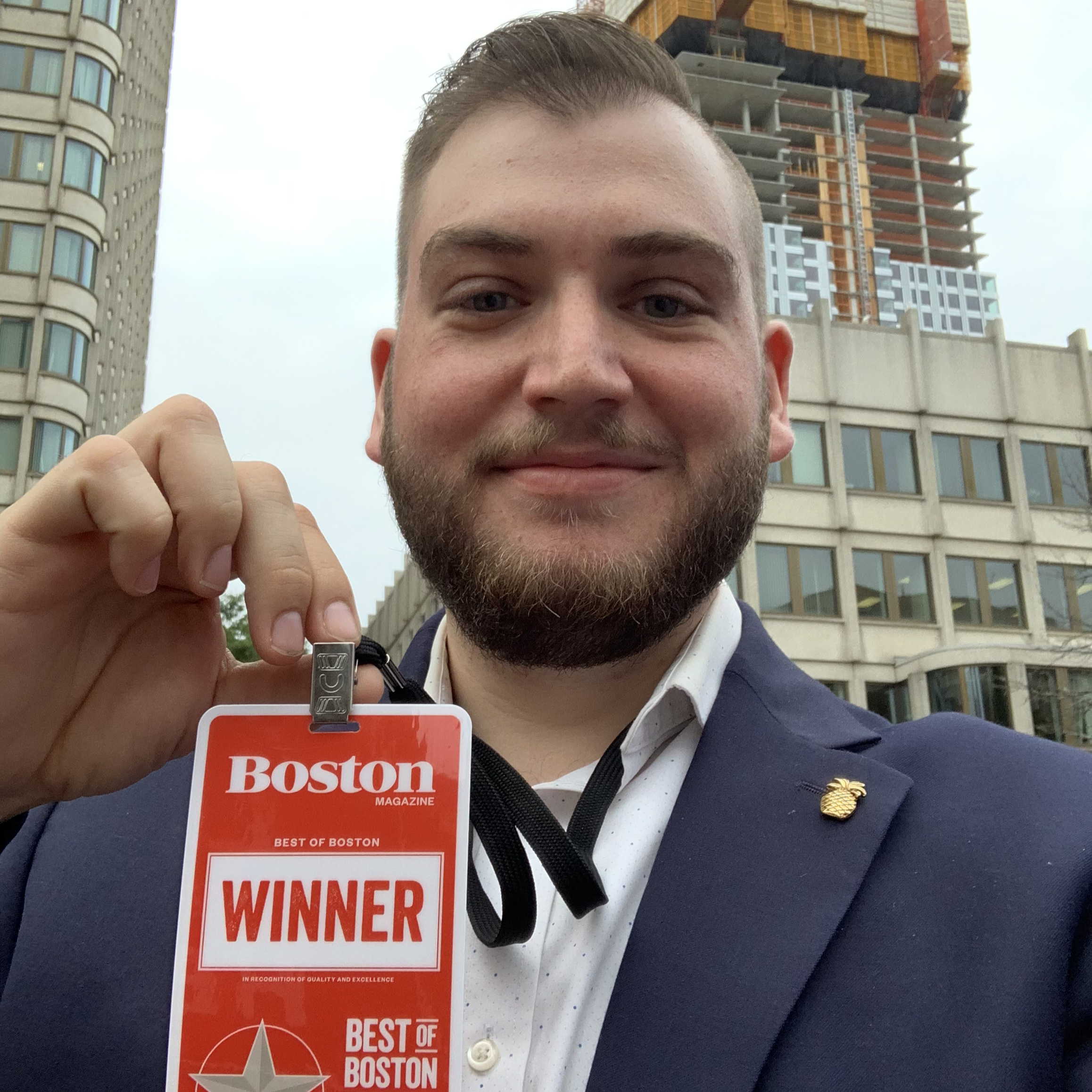 A man with a beard smiling and showing a badge that reads 'Winner' from Boston Magazine, standing outdoors in front of a cityscape with buildings and a construction site, dressed in a suit with a white shirt.