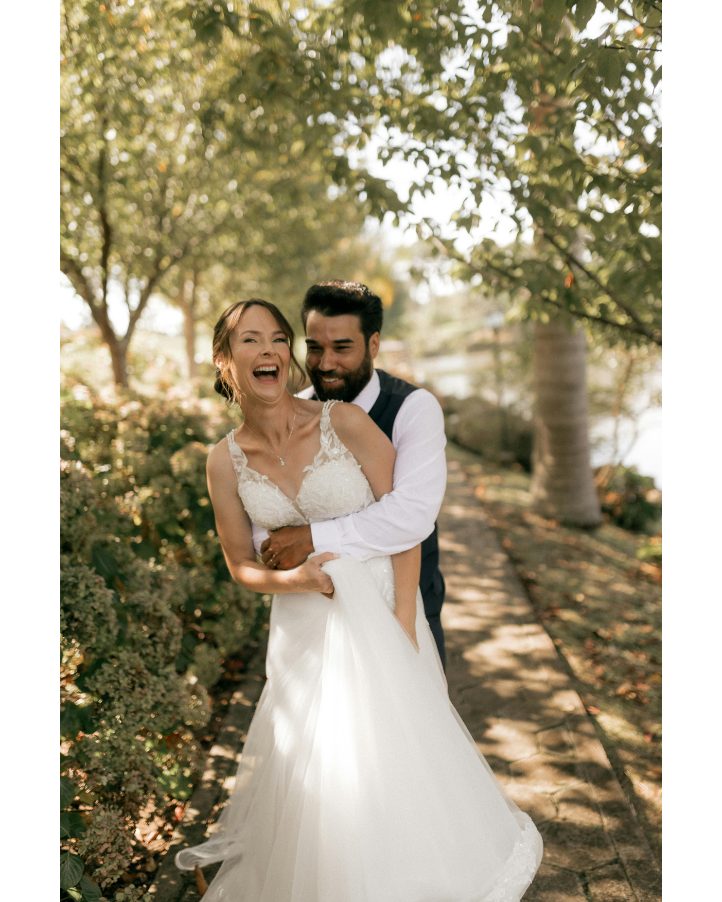 A bride and groom are smiling and laughing outdoors on a sunny day, with trees and greenery in the background. The bride is wearing a white wedding dress and the groom is wearing a dark vest and white shirt.