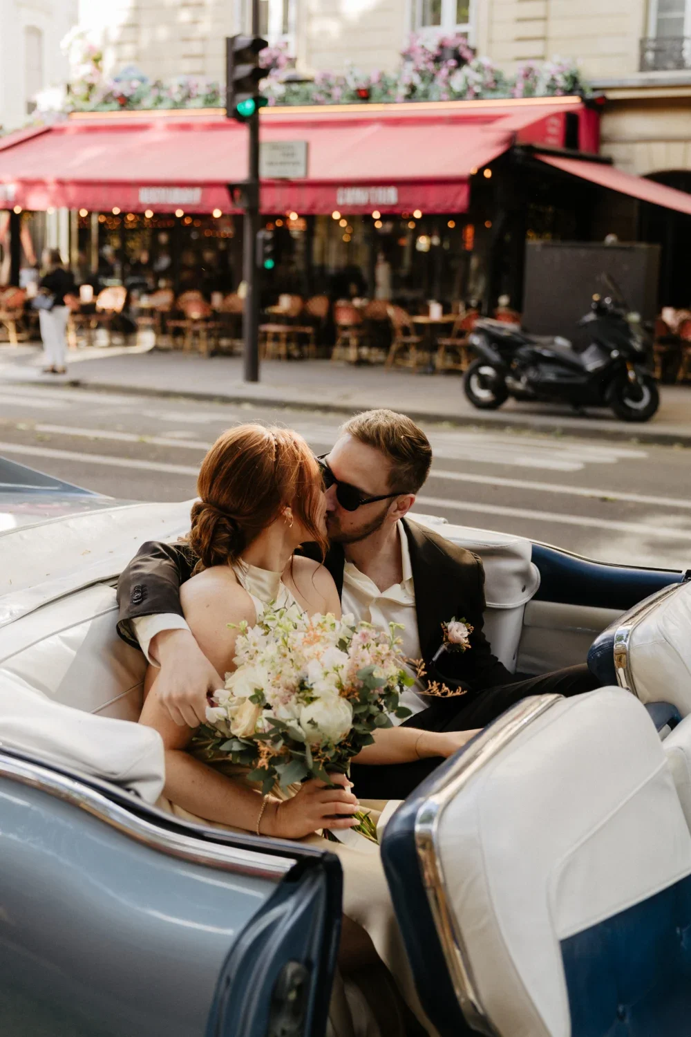 A newlywed couple kissing in a vintage car on a city street, with a restaurant and pedestrians in the background.