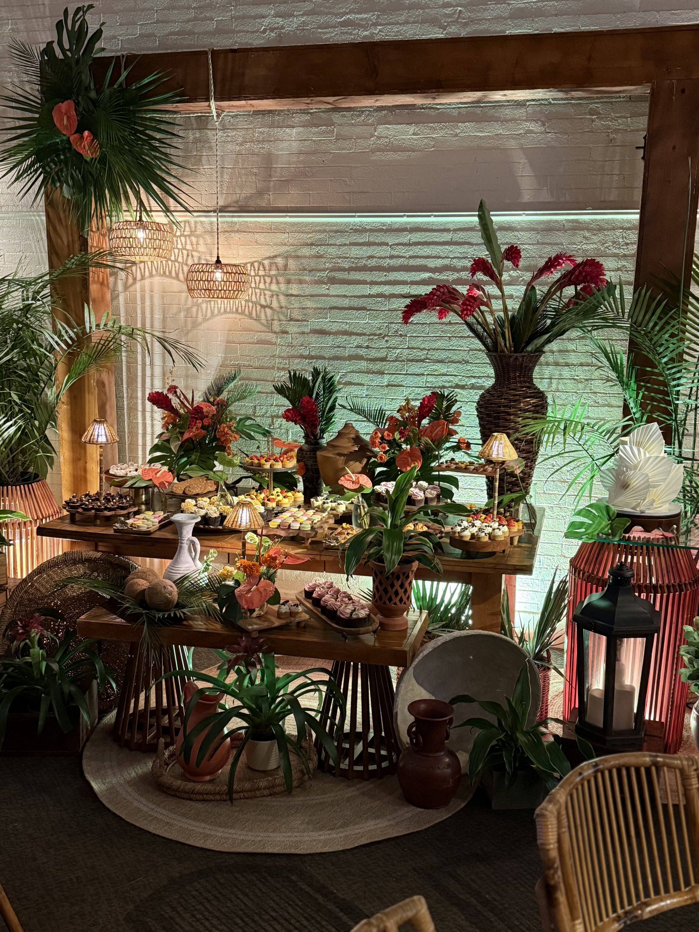 Decorative centerpiece table with desserts, surrounded by lush green plants, wicker furniture, and warm lighting in an indoor space.
