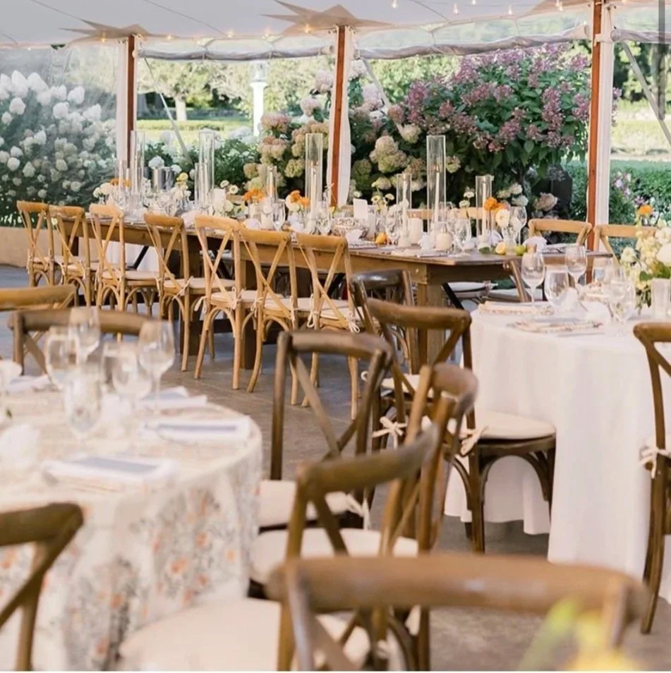 A decorated outdoor dining area with round and rectangular tables, wooden chairs, and floral centerpieces, set up for a wedding or event under a canopy with lot of pink and white flowers background.