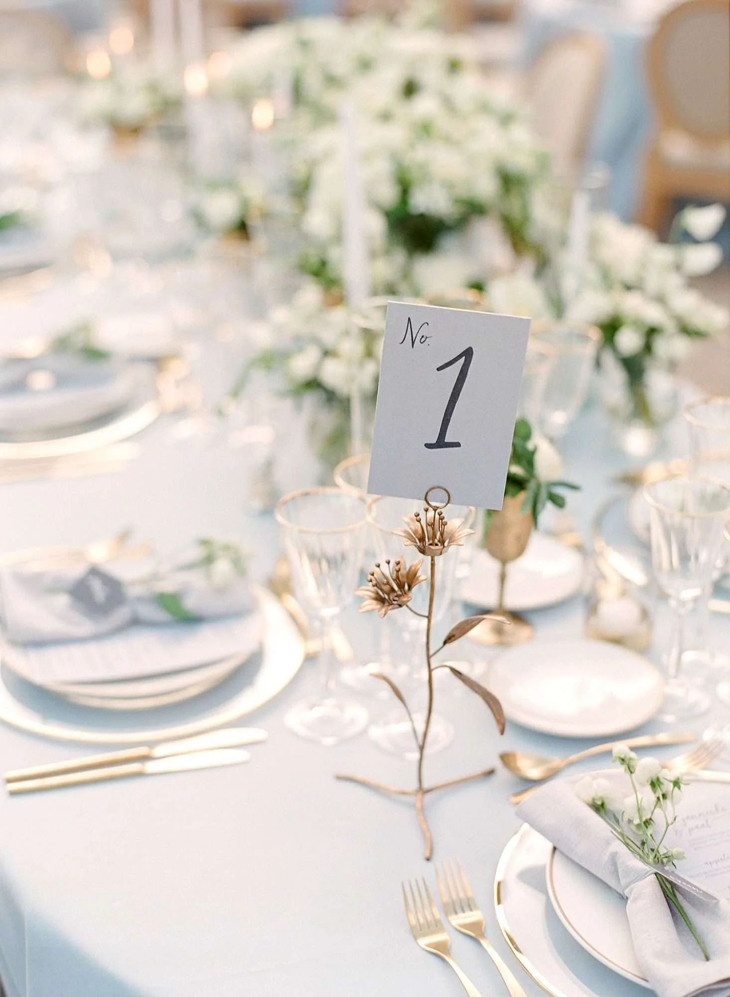 Elegant table setting with gold flatware, white plates, and floral centerpieces with a table number card labeled 'No. 1' in a gold holder. The table is decorated in a white and gold theme for a formal event or wedding reception.