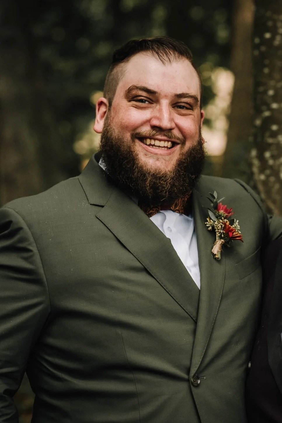Man with a beard smiling, dressed in a green suit with a boutonnière, outdoors with trees in the background.