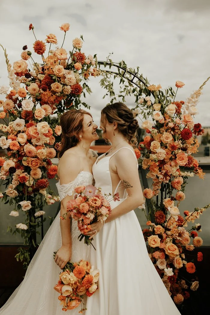 Two brides in wedding dresses smiling at each other in front of a large floral arch with pink, peach, and red flowers.