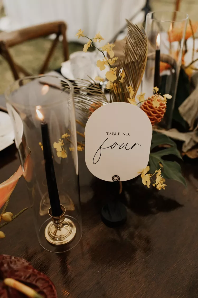 Table setting with a white table number card labeled 'Table No. Four,' surrounded by two tall glass candle holders with lit black candles, decorative dried leaves, yellow flowers, and foliage on a dark wooden table.