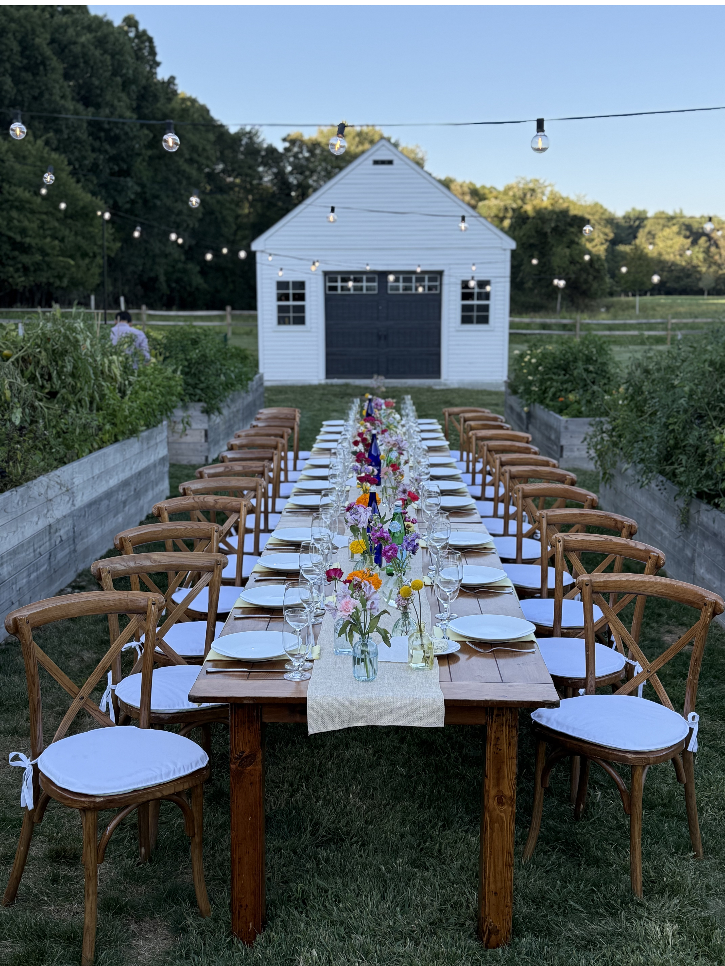 Long outdoor dining table with white table runners, floral centerpieces, glassware, and wooden chairs, set up in a backyard with string lights, greenery, and a white barn in the background.