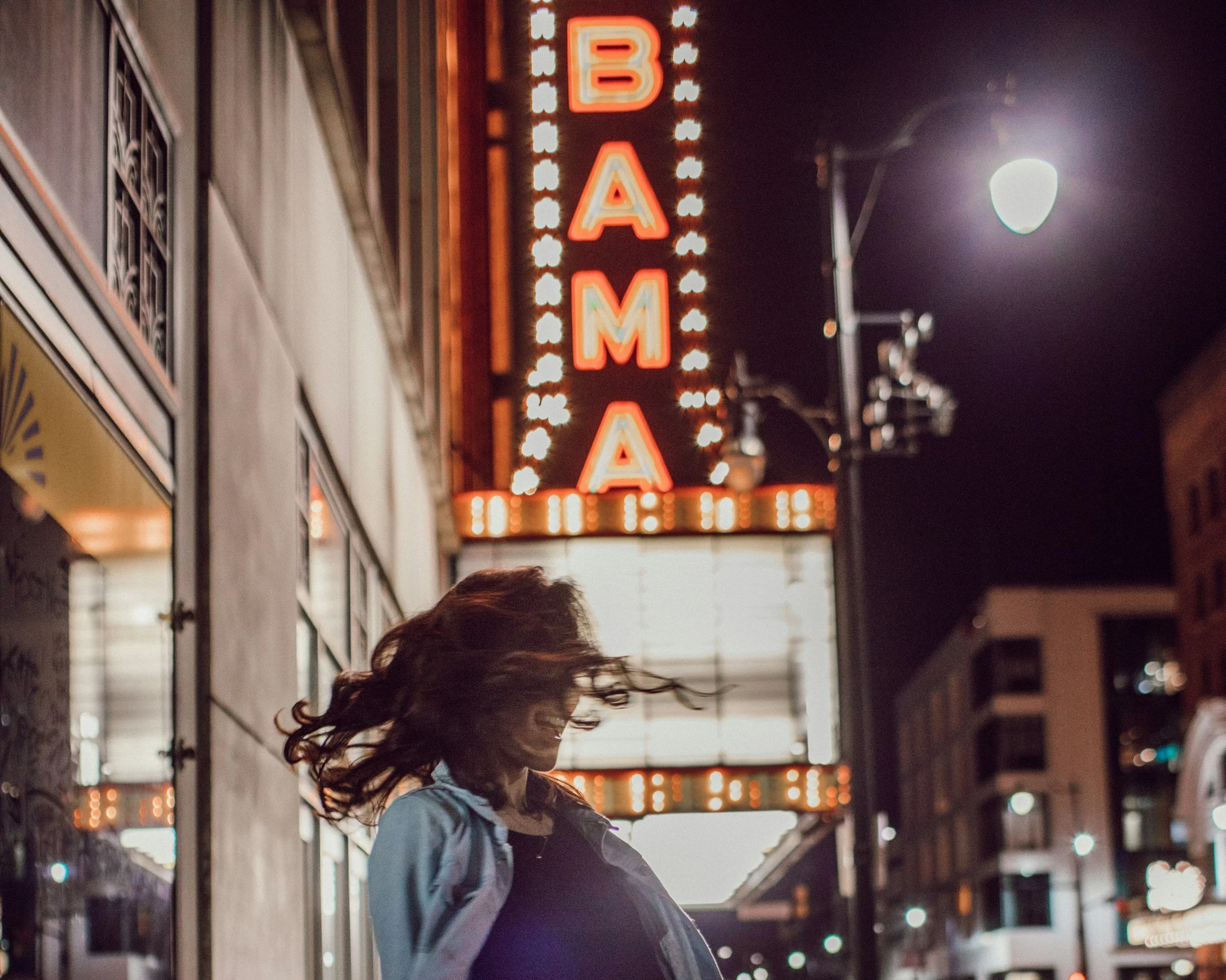 Nighttime street scene with a person with long hair and a gray jacket, walking past a building with illuminated signs, including a large vertical neon sign that reads 'BAMA', with bright lights outlining the sign.