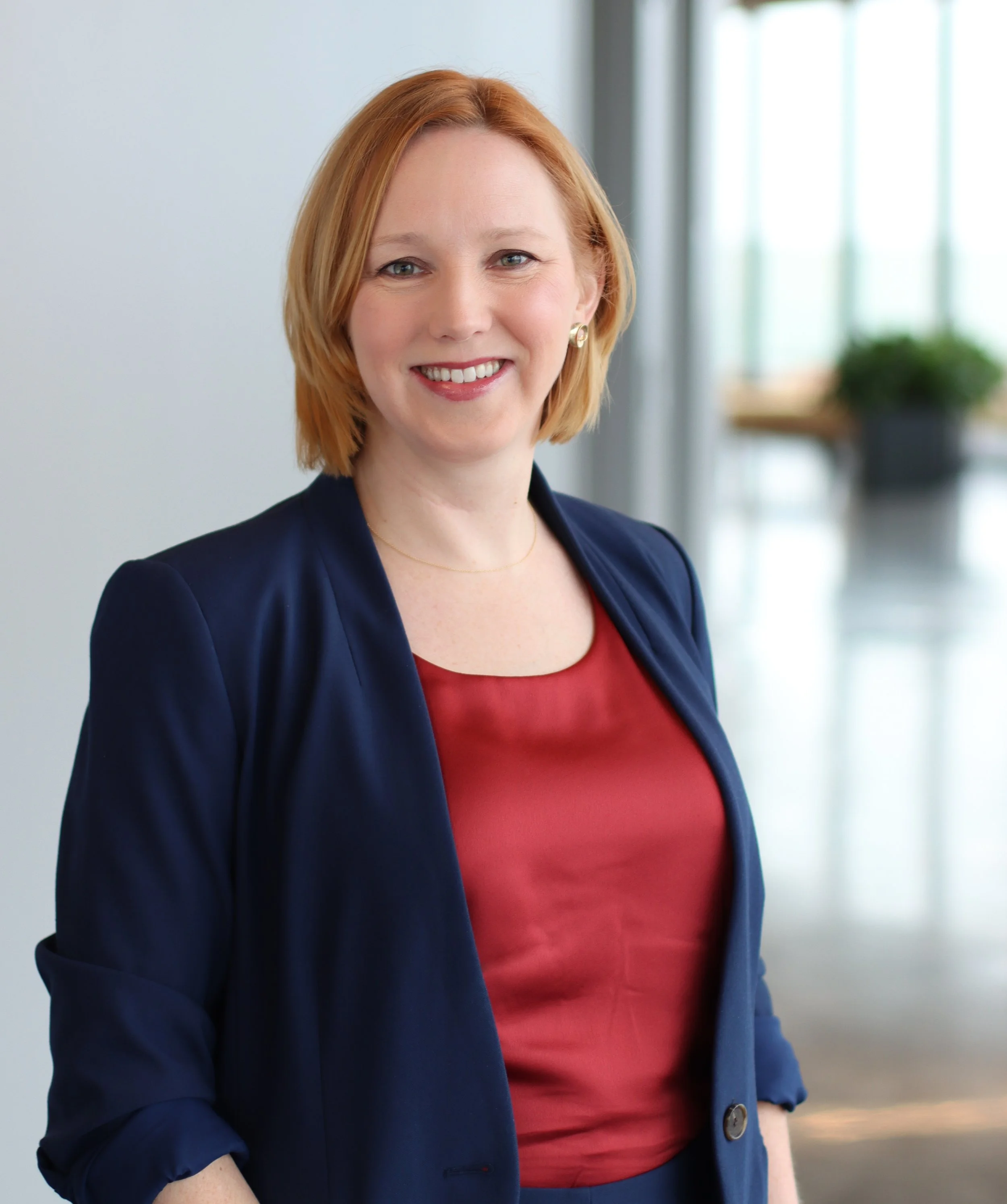 Close-up of a woman with short red hair, wearing a navy blazer and a red top, smiling indoors with a blurred background featuring large windows and a potted plant.