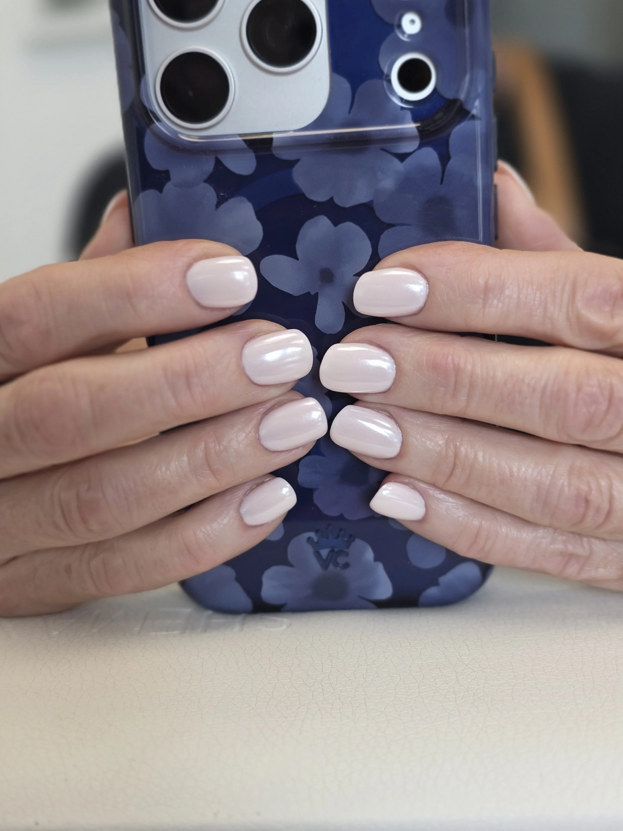Person holding a smartphone with a floral blue and white patterned case. The person has light-colored painted nails and is seated at a table.