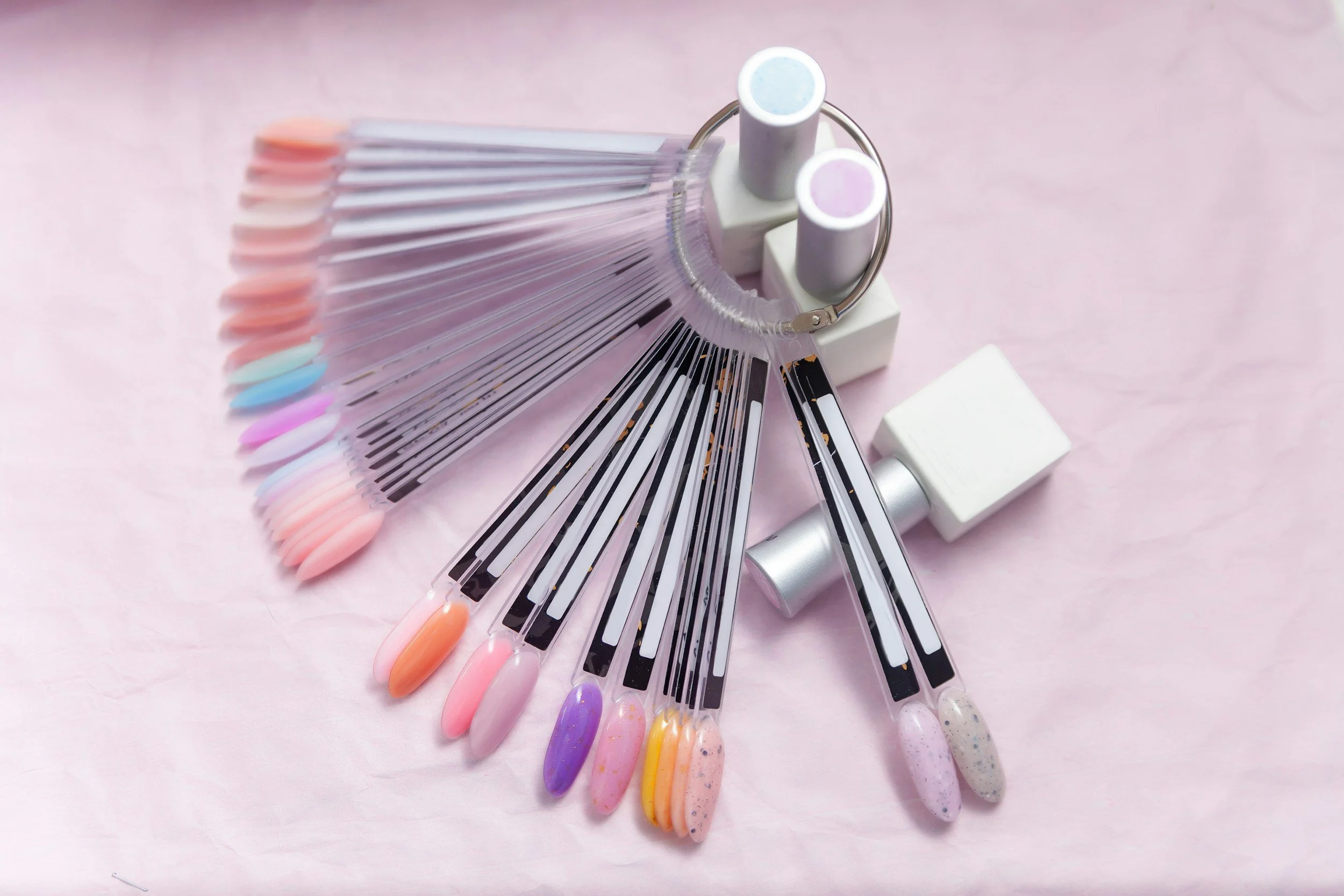 Display of various nail polish swatches on nail tips, fanned out next to a nail UV lamp and nail gel polish bottles, set on a pink background.