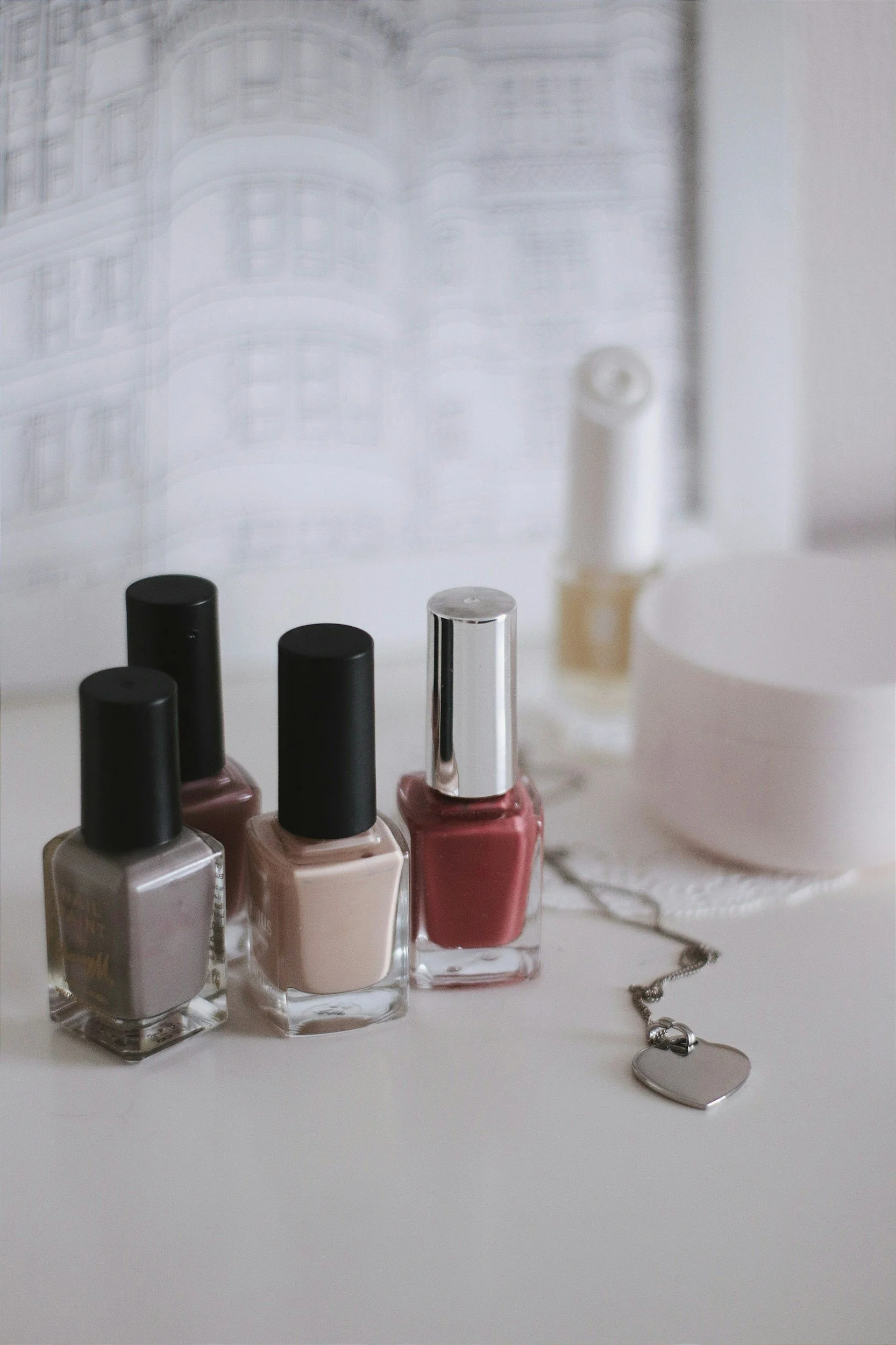 Four bottles of nail polish in shades of beige, pink, red, and taupe, a silver necklace with a heart-shaped pendant, a white round container, and a white skincare or cosmetic spray bottle, all on a white surface with a soft background.
