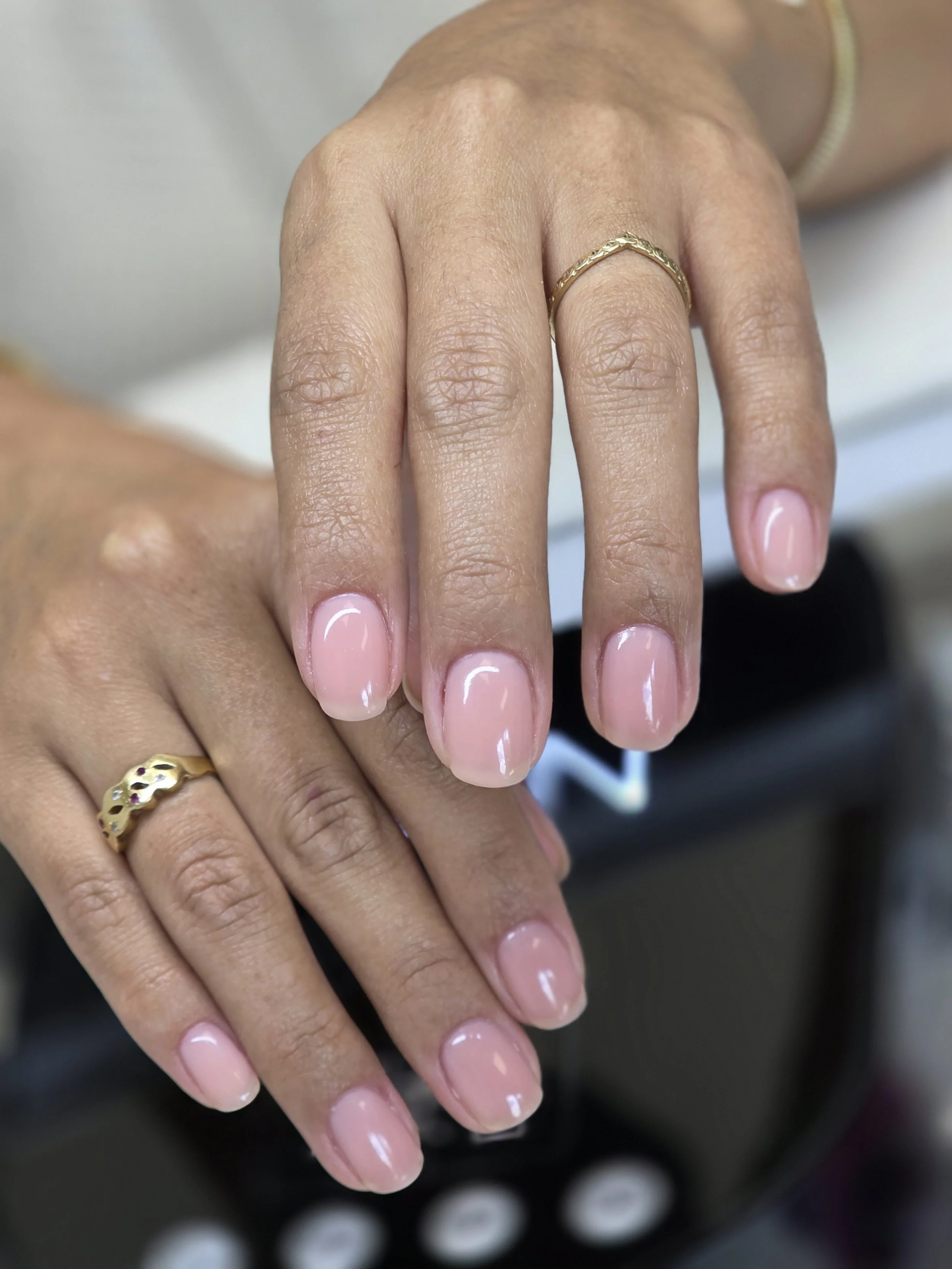 Close-up of a hand with neatly manicured nails painted in a light pink color, showcasing a gold ring on the middle finger and a gold bracelet on the wrist.
